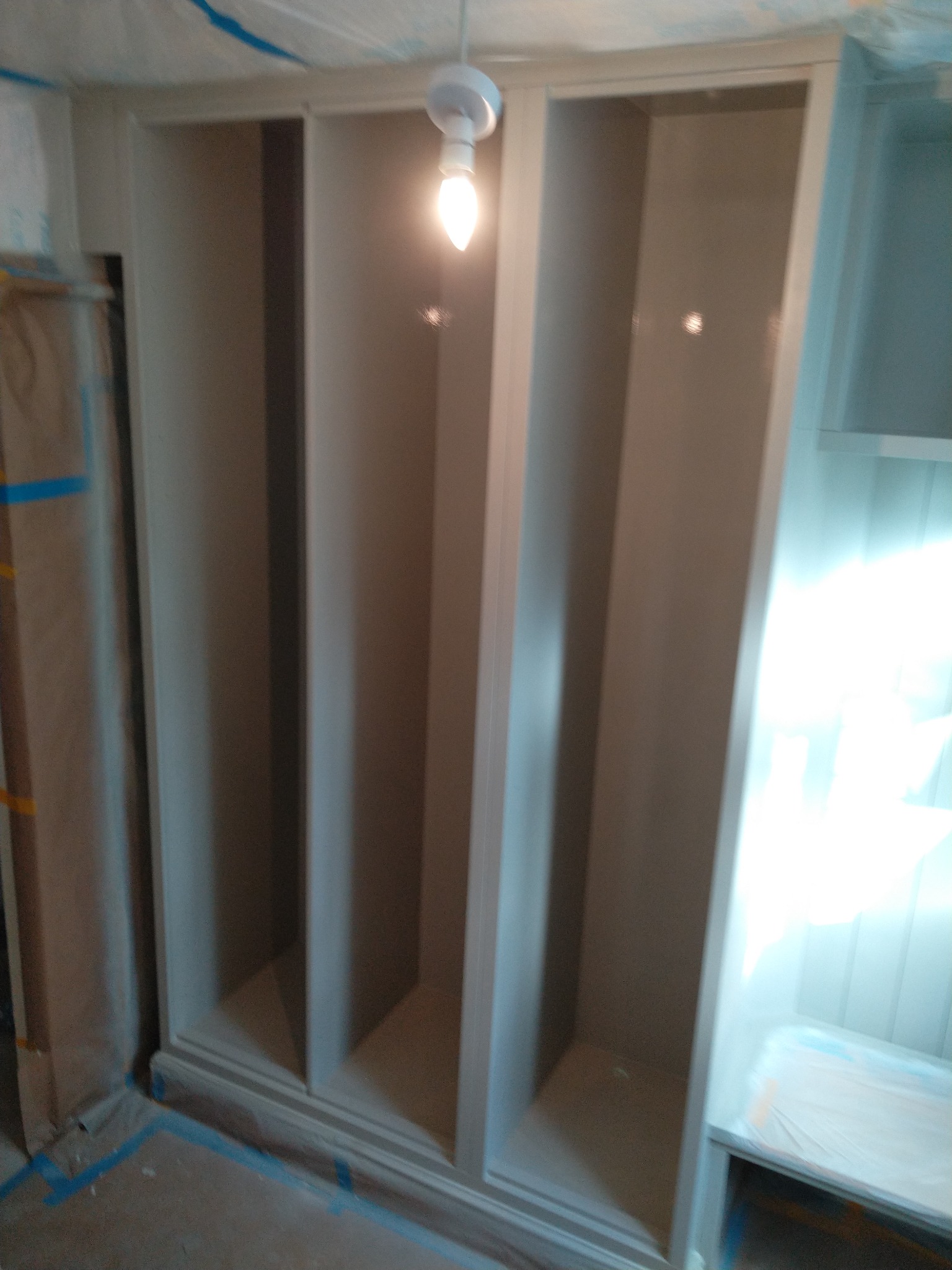 Wnętrze szafy wnękowej z trzema pustymi przegródkami, świeżo pomalowana na szaro, z widocznymi śladami taśmy malarskiej na podłodze i zabezpieczeniem folią malarską sufitu.