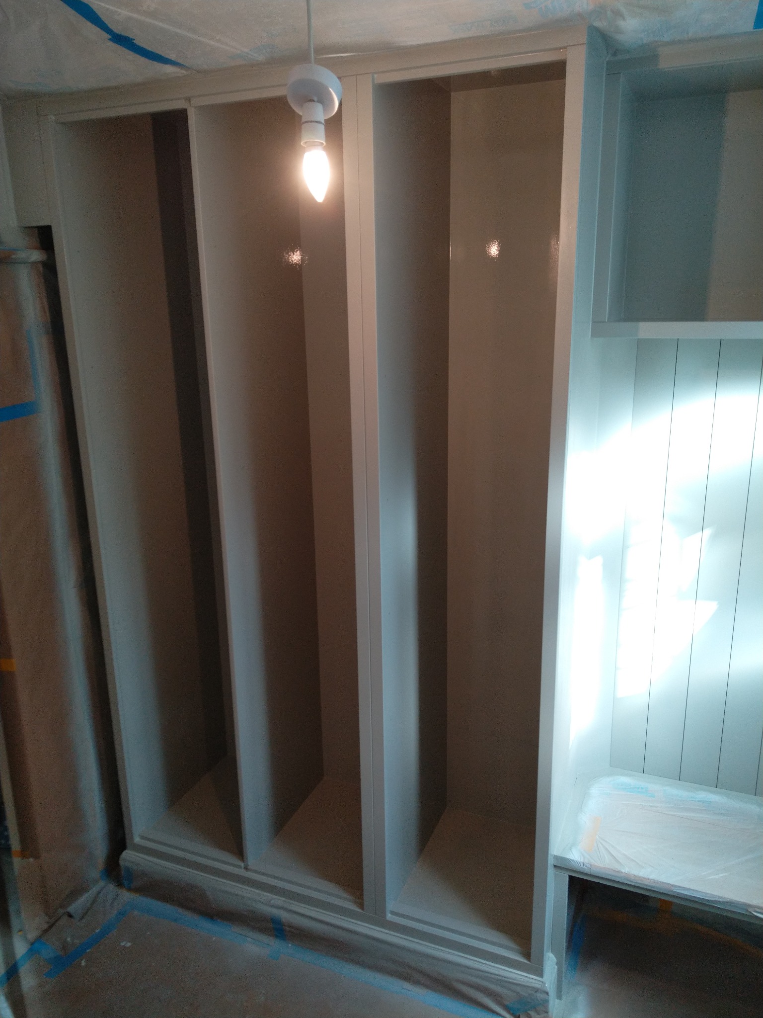 Pomalowana na szaro zabudowa meblowa w trakcie prac wykończeniowych, zabezpieczona folią malarską, widoczna żarówka