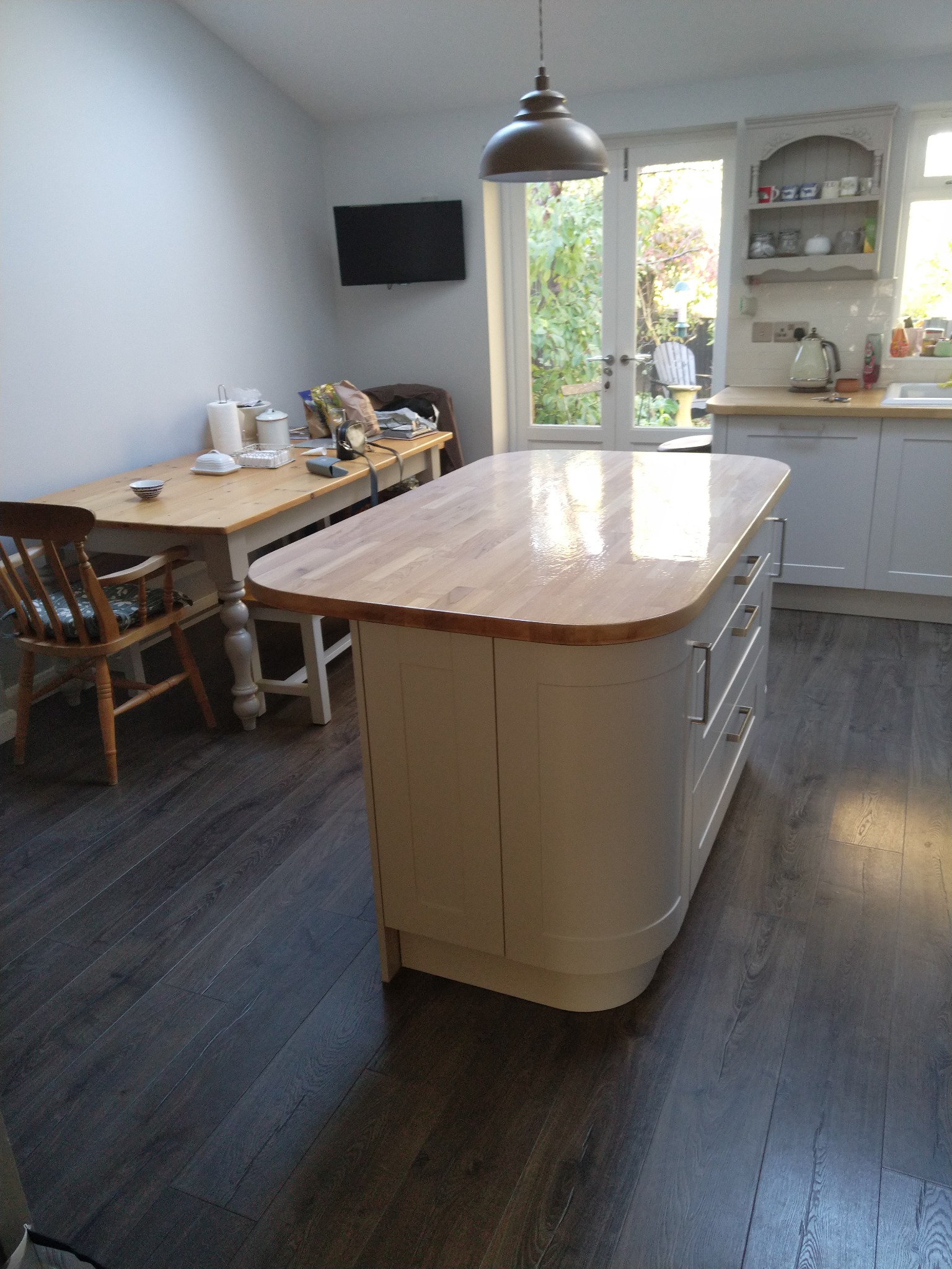 Wnętrze kuchni po remoncie: wyspa kuchenna z drewnianym blatem i szarymi frontami, widok na stół z krzesłami i okno z widokiem na ogród.