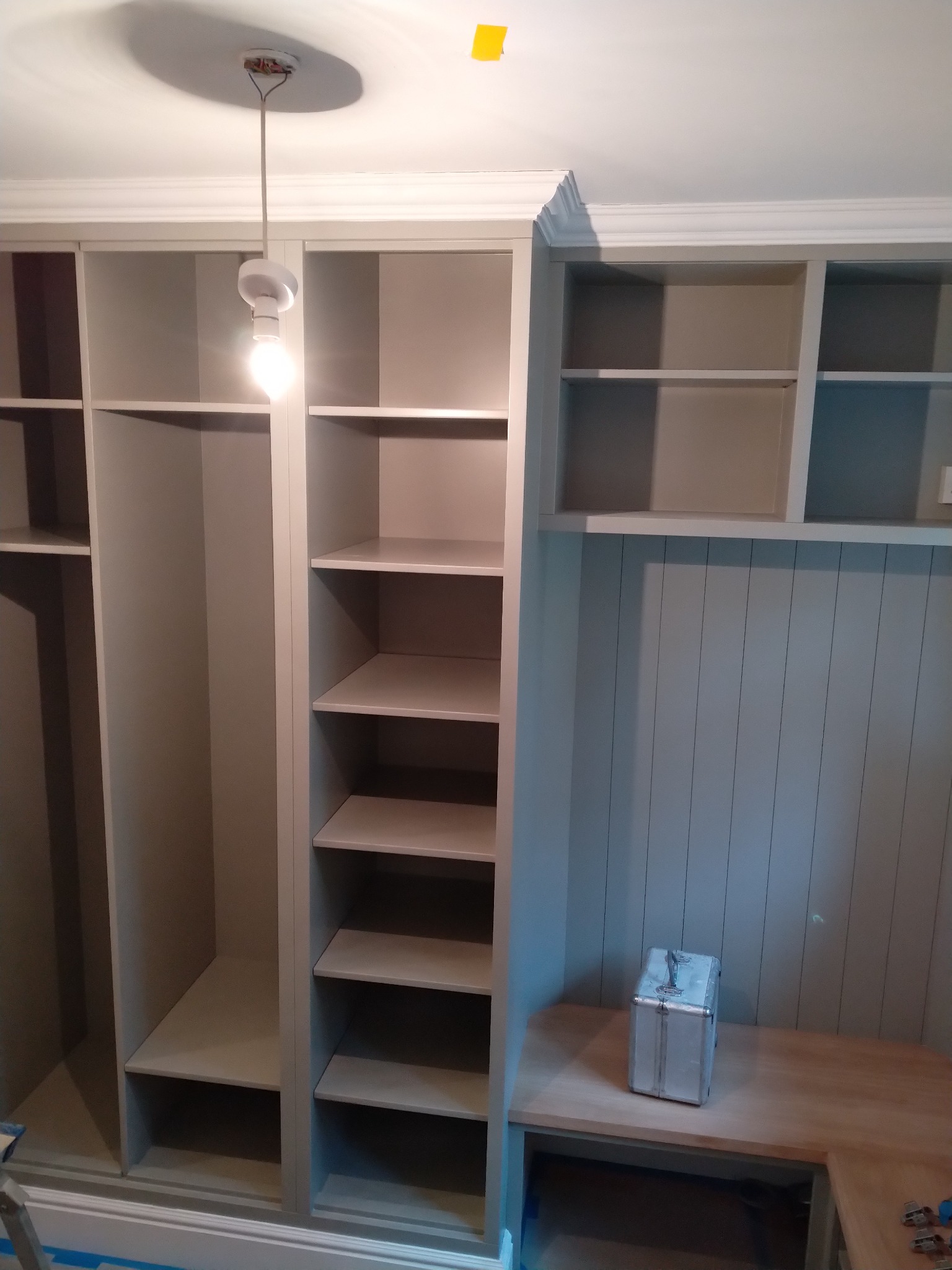 Szare regały z półkami i wnękami, biała listwa przypodłogowa i sufitowa, wisząca żarówka, aluminiowa skrzynka na blacie, fragment drabiny malarskiej w lewym dolnym rogu.