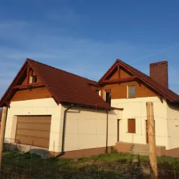 Nowo wybudowany dom jednorodzinny z brązową dachówką, kremową elewacją i drewnianymi elementami wykończeniowymi, sfotografowany zza ogrodzenia z siatki.