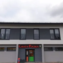 Dwukondygnacyjny budynek z szarą elewacją i ciemnoszarymi panelami na górnej kondygnacji, z dużym czerwonym napisem 'CHATA POLSKA' nad wejściem i kolorowymi drzwiami.