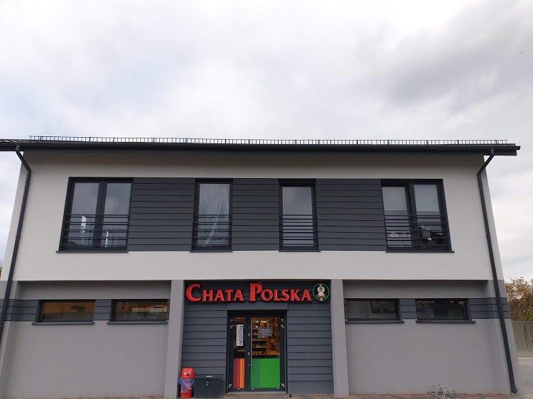 Dwukondygnacyjny budynek z szarą elewacją i ciemnoszarymi panelami na górnej kondygnacji, z dużym czerwonym napisem 'CHATA POLSKA' nad wejściem i kolorowymi drzwiami.