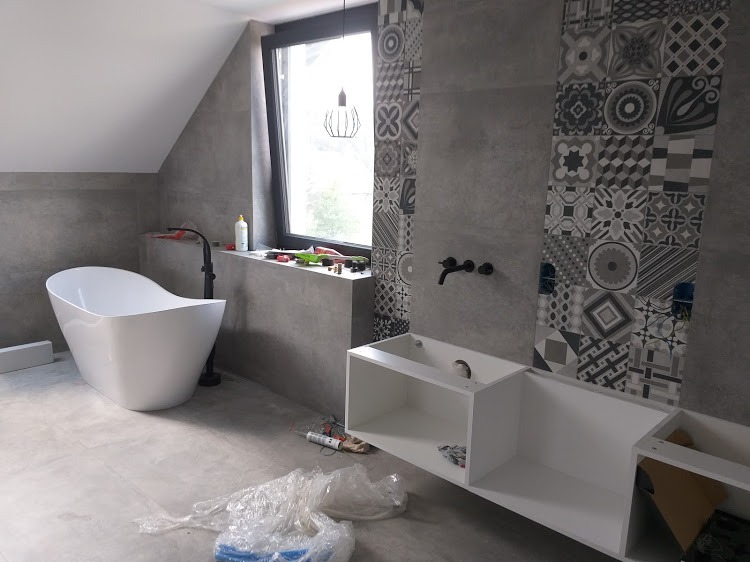 Łazienka w trakcie wykańczania: wolnostojąca wanna, szara ściana z wzorzystymi płytkami, biała zabudowa pod umywalkę, widoczne narzędzia i folia ochronna.