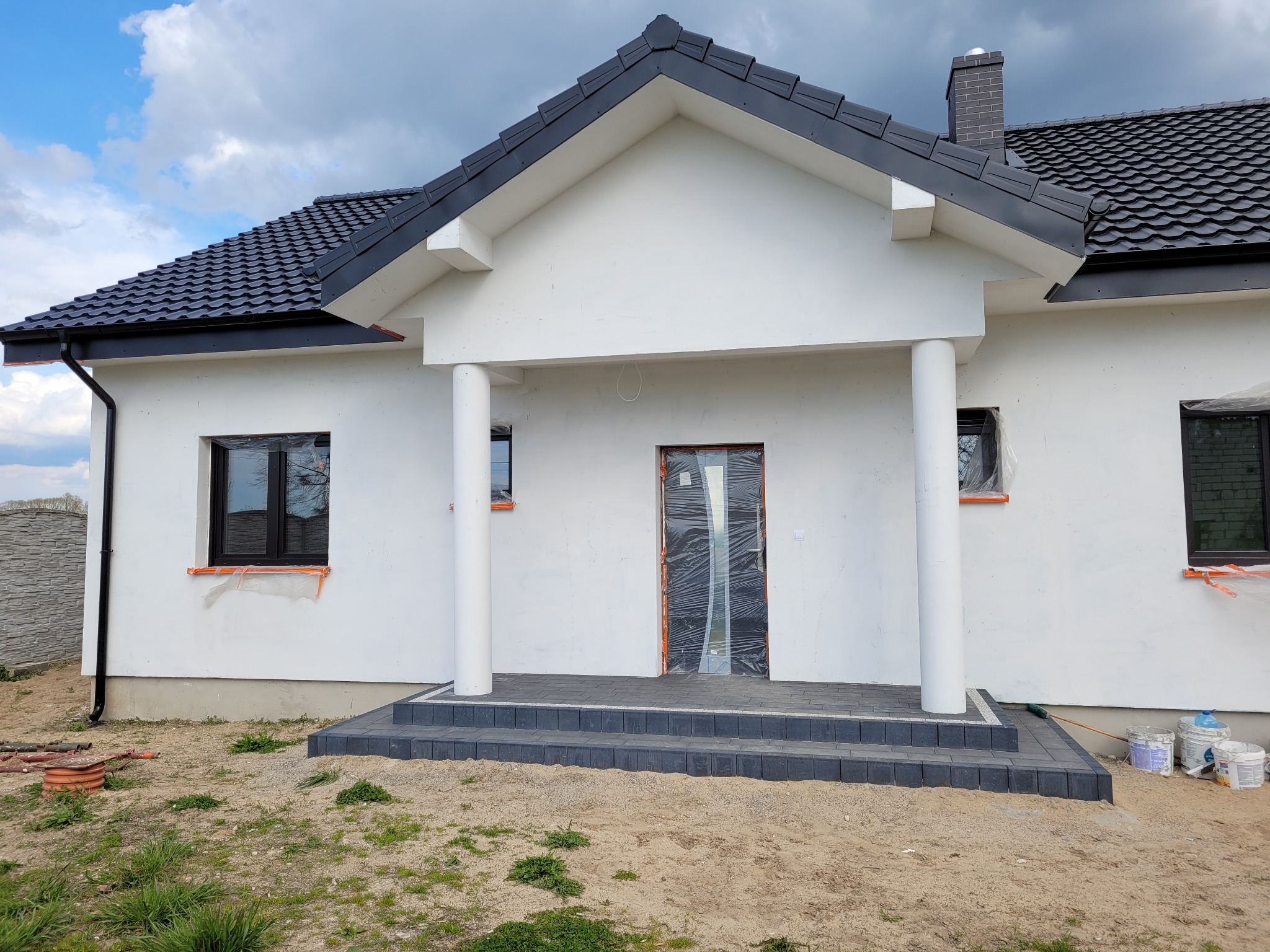 Niedawno wybudowany dom jednorodzinny w Chodzieży z białymi ścianami zewnętrznymi, ciemną dachówką i wykończonym wejściem z szarej kostki brukowej. Okna i drzwi zabezpieczone folią ochronną.