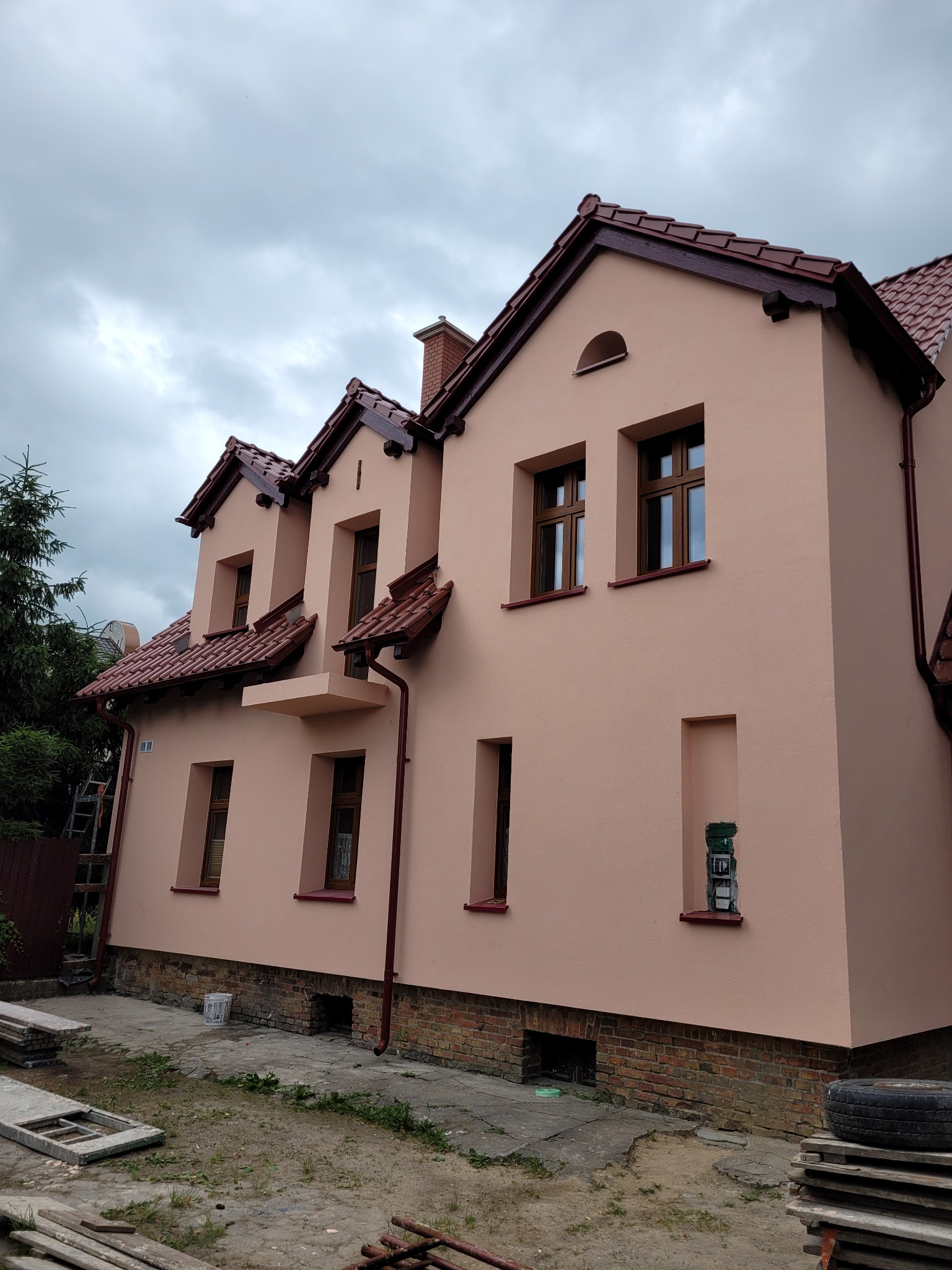 Dwupiętrowy dom z elewacją w odcieniach beżu i brązu, widoczny z zewnątrz, z elementami wykończeniowymi i fragmentem terenu przed budynkiem.