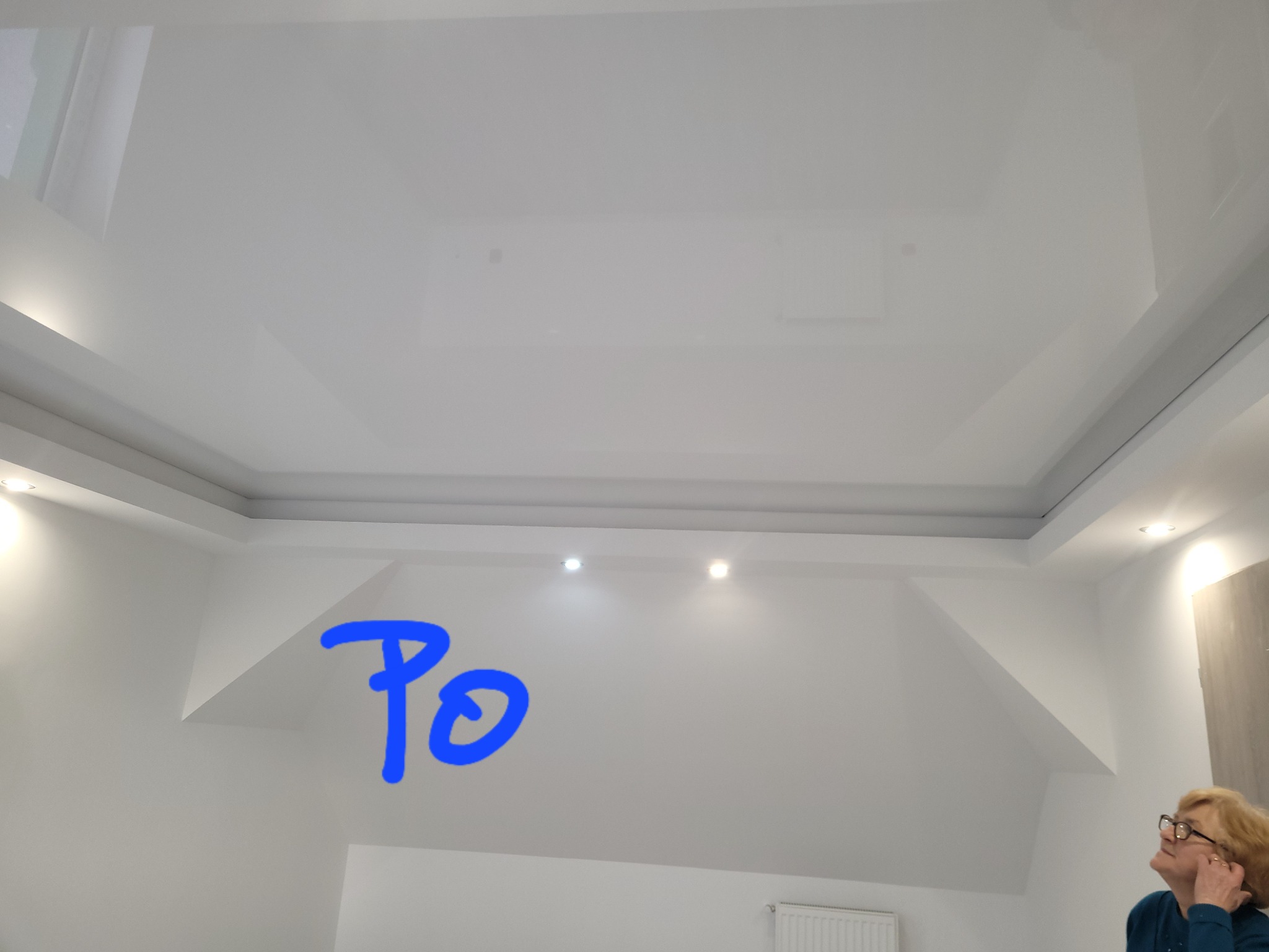 Biały, błyszczący sufit napinany z oświetleniem punktowym, widoczny fragment ściany i osoba w rogu kadru, napis 'Po' na ścianie.