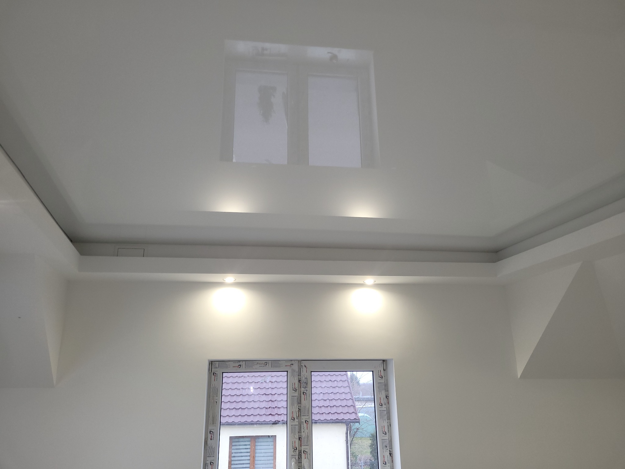 Białe ściany i sufit z wnękami, oświetleniem punktowym i widokiem na dom z czerwonym dachem przez okno.