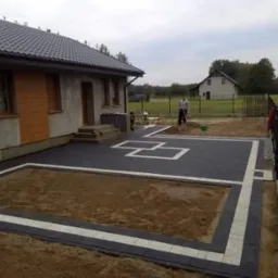 Częściowo ułożona kostka brukowa w kolorze grafitowym z białymi akcentami na podjeździe przed domem, widoczny fragment ogrodu i pracownik z łopatą w tle.