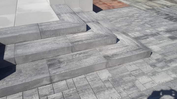 Schody wejściowe wyłożone szarą kostką brukową o nieregularnym wzorze, z widocznym cieniem rzucanym przez fotografa