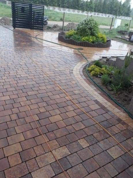 Świeżo ułożona, mokra kostka brukowa w odcieniach brązu, z dekoracyjnym obramowaniem z jaśniejszych kostek, widoczny fragment czarnego ogrodzenia i rabata z roślinami w tle, na kostce leży...