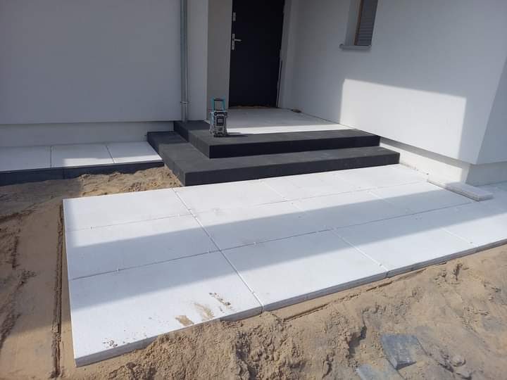 Biała kostka brukowa ułożona przed wejściem do domu, z czarnymi schodami i laserowym niwelatorem na jednym ze stopni.