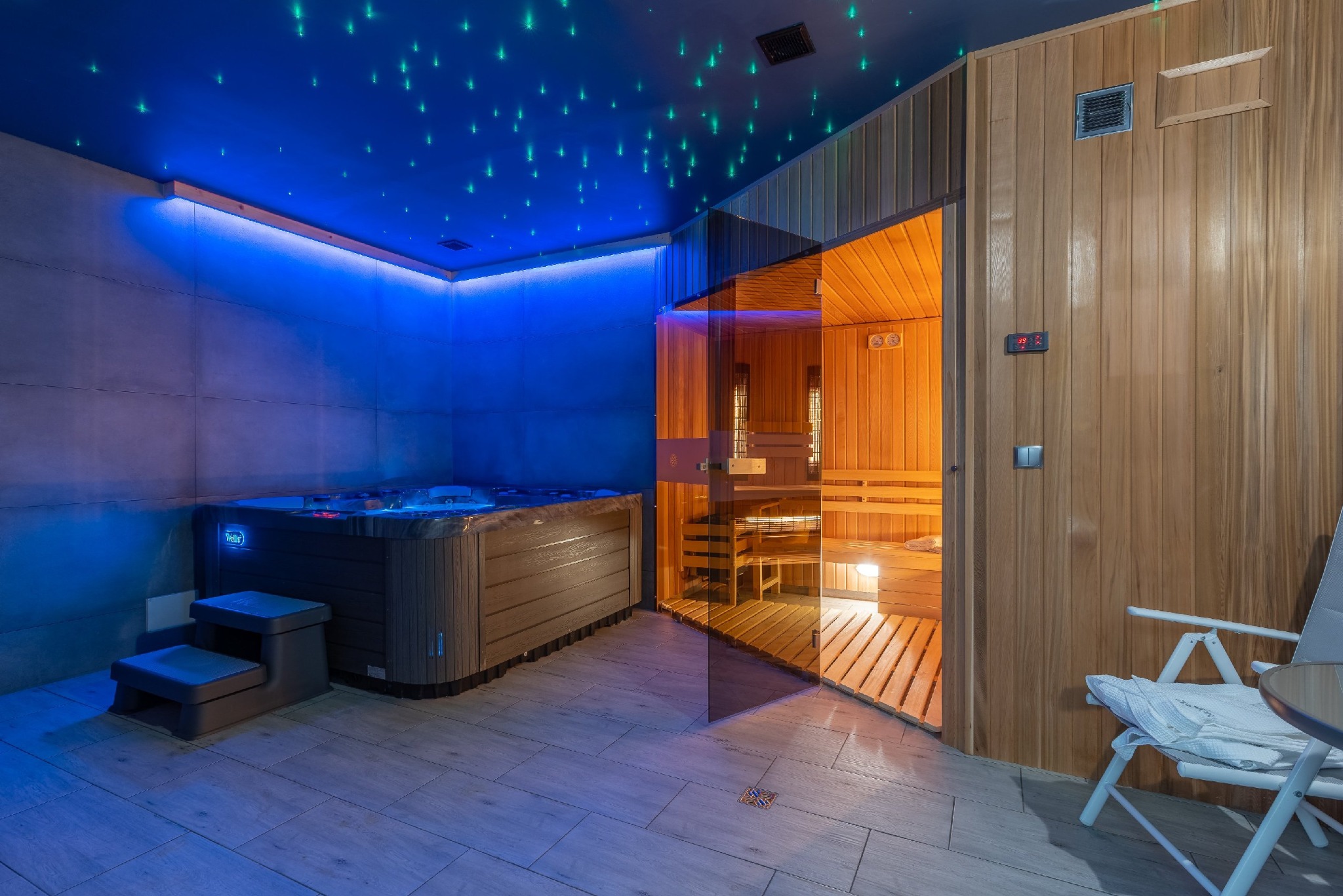 Nowoczesne wnętrze spa z drewnianą sauną i jacuzzi podświetlonym na niebiesko, sufit z dekoracyjnym oświetleniem imitującym gwiazdy, płytki podłogowe w odcieniach szarości i białe krzesło...