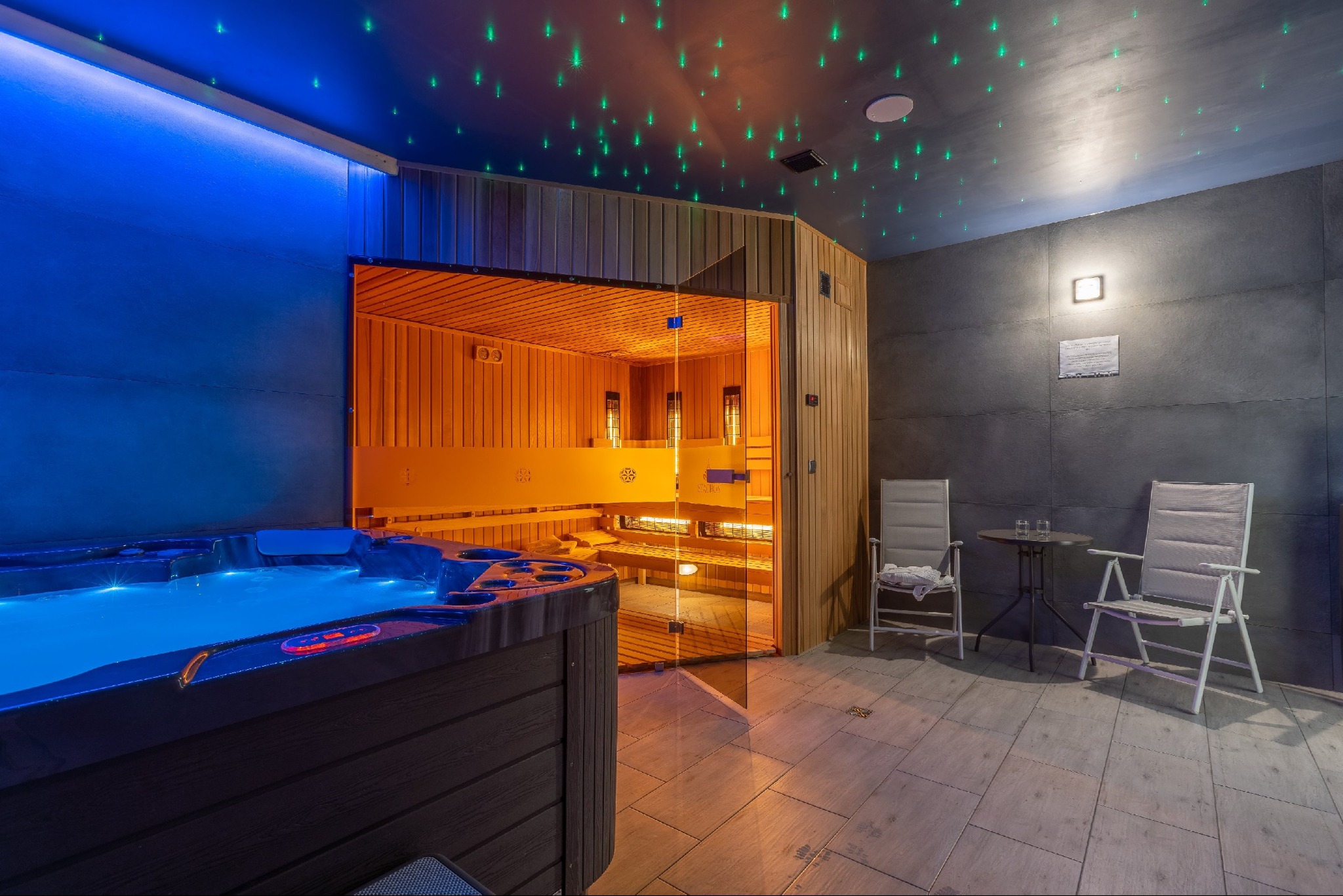 Nowoczesne wnętrze strefy relaksu z podświetlanym jacuzzi, sauną z przeszklonymi drzwiami, oraz strefą wypoczynkową z krzesłami i stolikiem, oświetlone dekoracyjnymi lampkami w suficie.