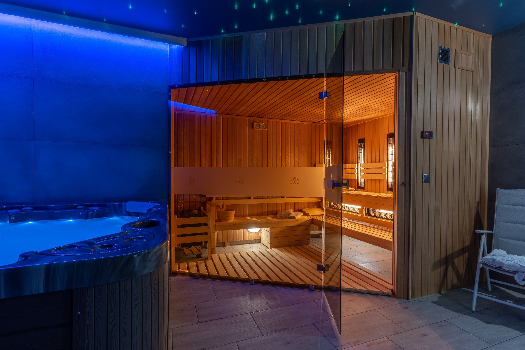 Nowoczesna strefa relaksu z otwartą sauną fińską i jacuzzi podświetlonym na niebiesko, sufit z dekoracyjnym oświetleniem punktowym.