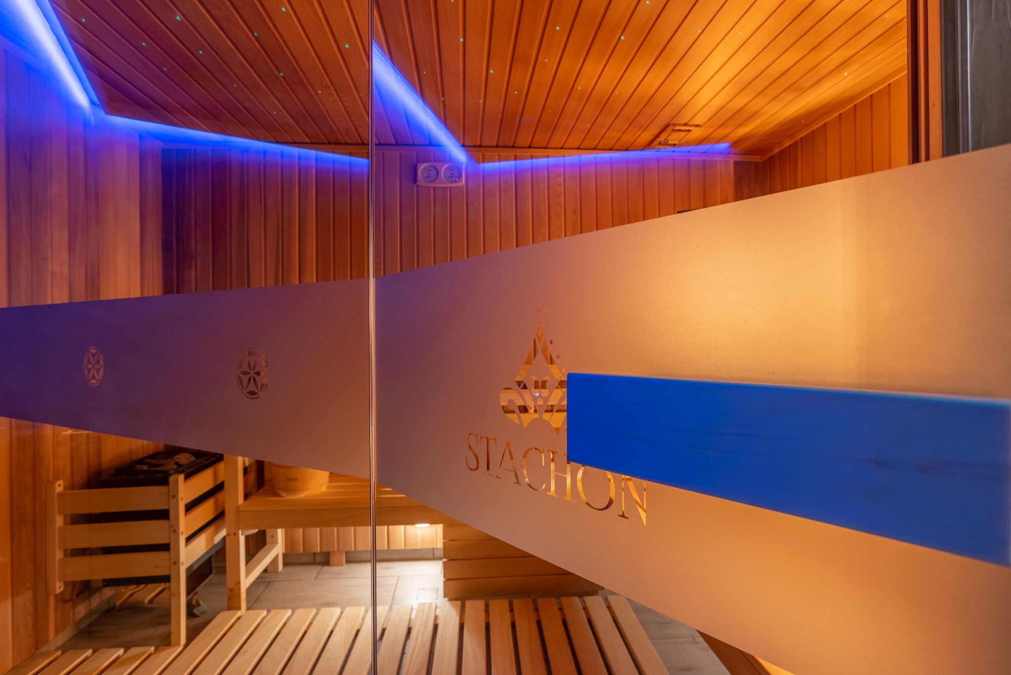 Wnętrze sauny z drewnianymi ławkami, piecem i podświetleniem LED, widoczne przez matową szybę z logo firmy Stachon.