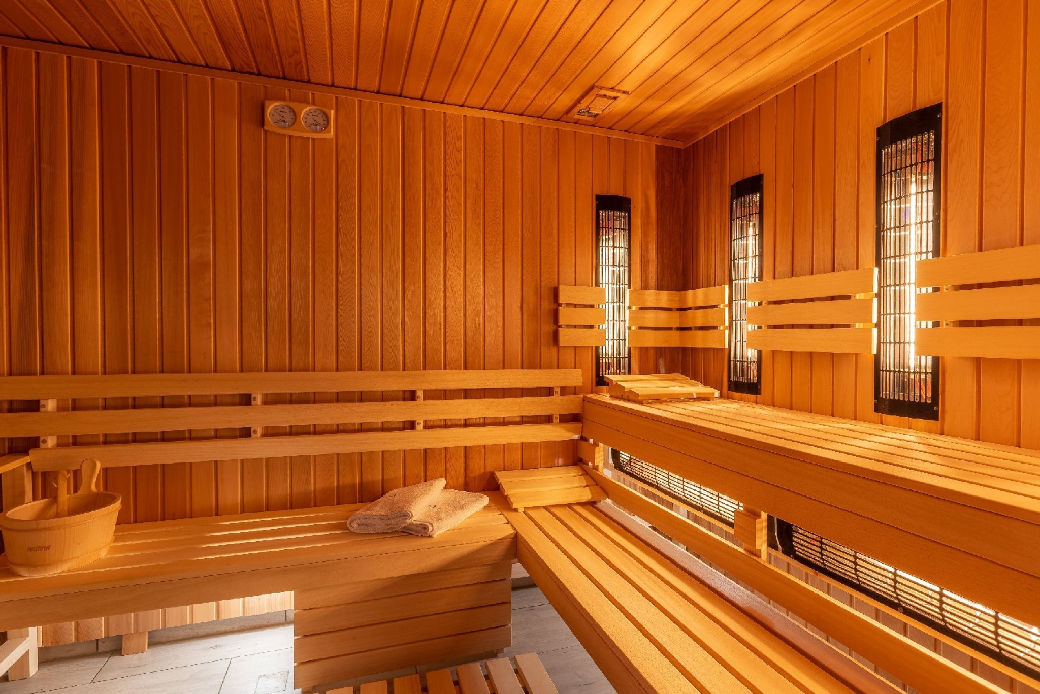 Wnętrze sauny wykończone drewnem, z drewnianymi ławkami, oświetleniem halogenowym i akcesoriami w postaci drewnianego wiadra i ręczników.