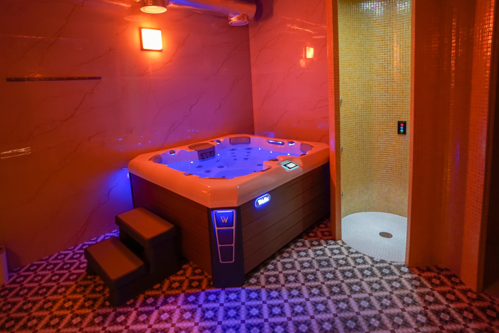 Nowoczesna wanna z hydromasażem z podświetleniem LED w pomieszczeniu z mozaikową podłogą i kabiną prysznicową wyłożoną drobną, żółtą mozaiką.