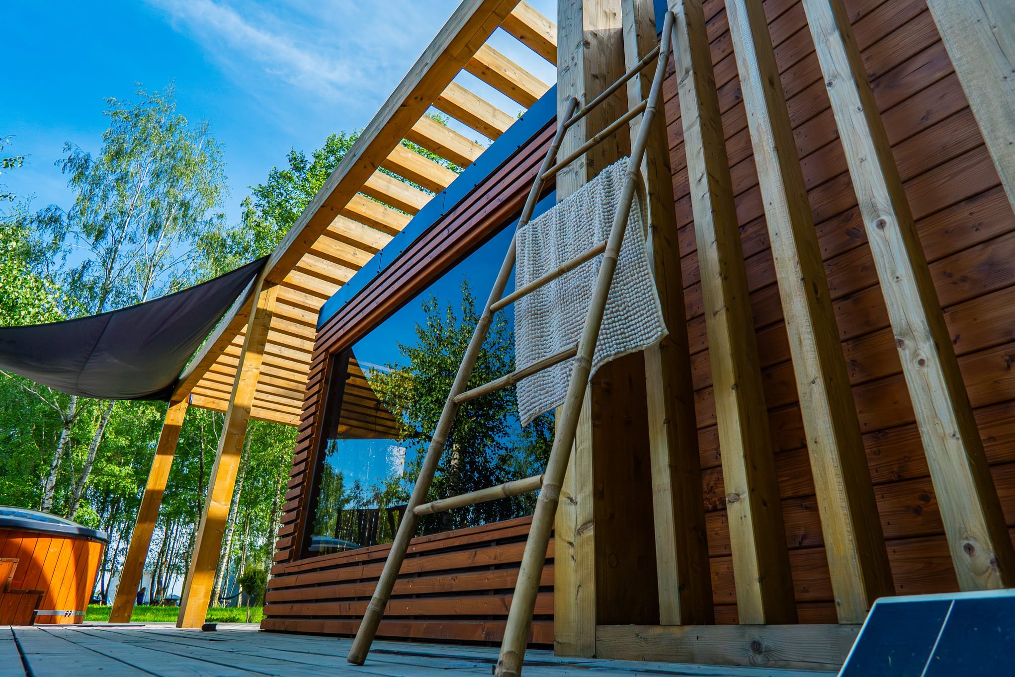 Drewniana sauna z dużym oknem, bambusową drabinką i ręcznikiem na zewnątrz, na tle zieleni i błękitnego nieba.