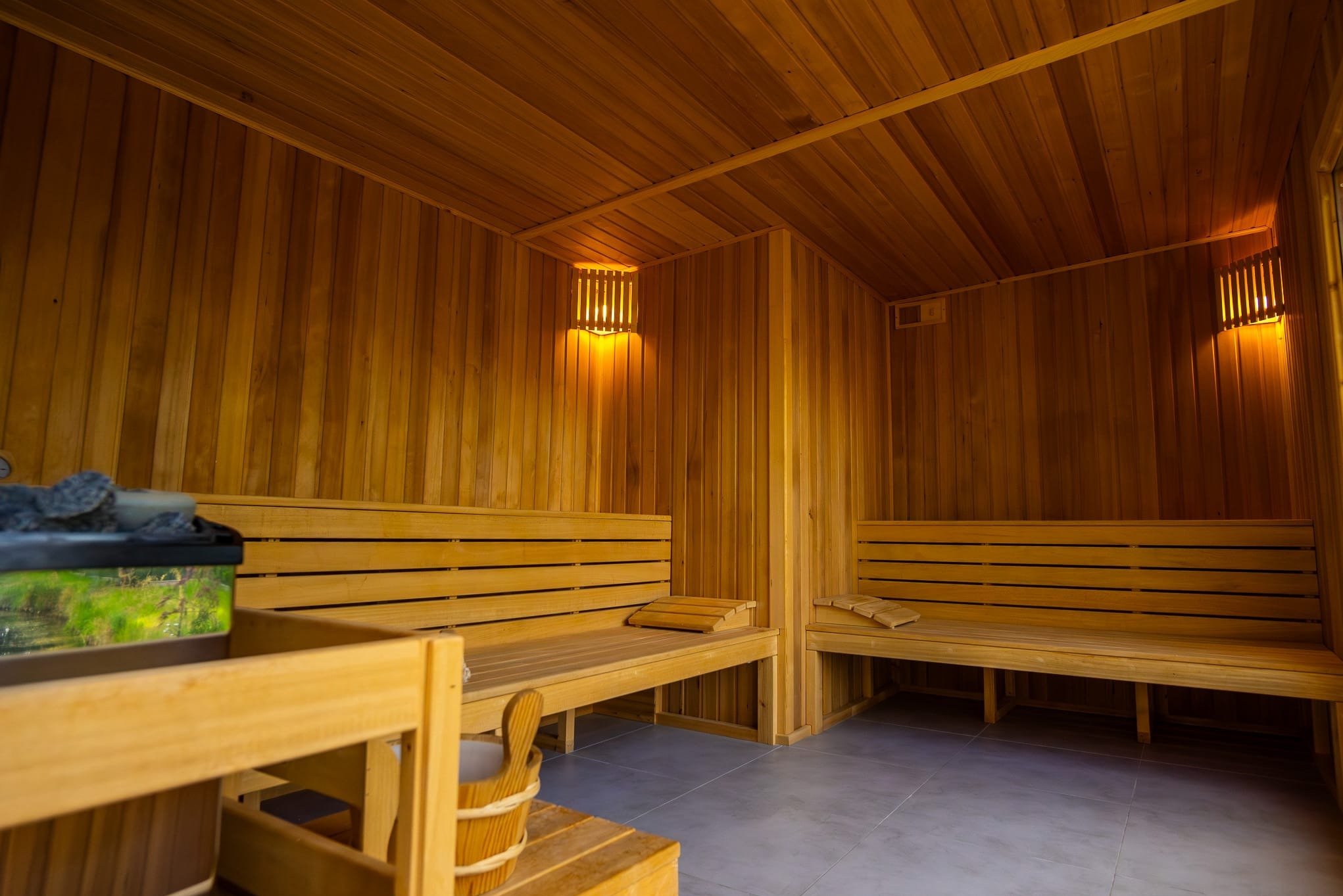 Wnętrze sauny wykończone drewnem z podświetlanymi lampami ściennymi, z piecem z kamieniami i dekoracyjnym panelem z motywem krajobrazu.