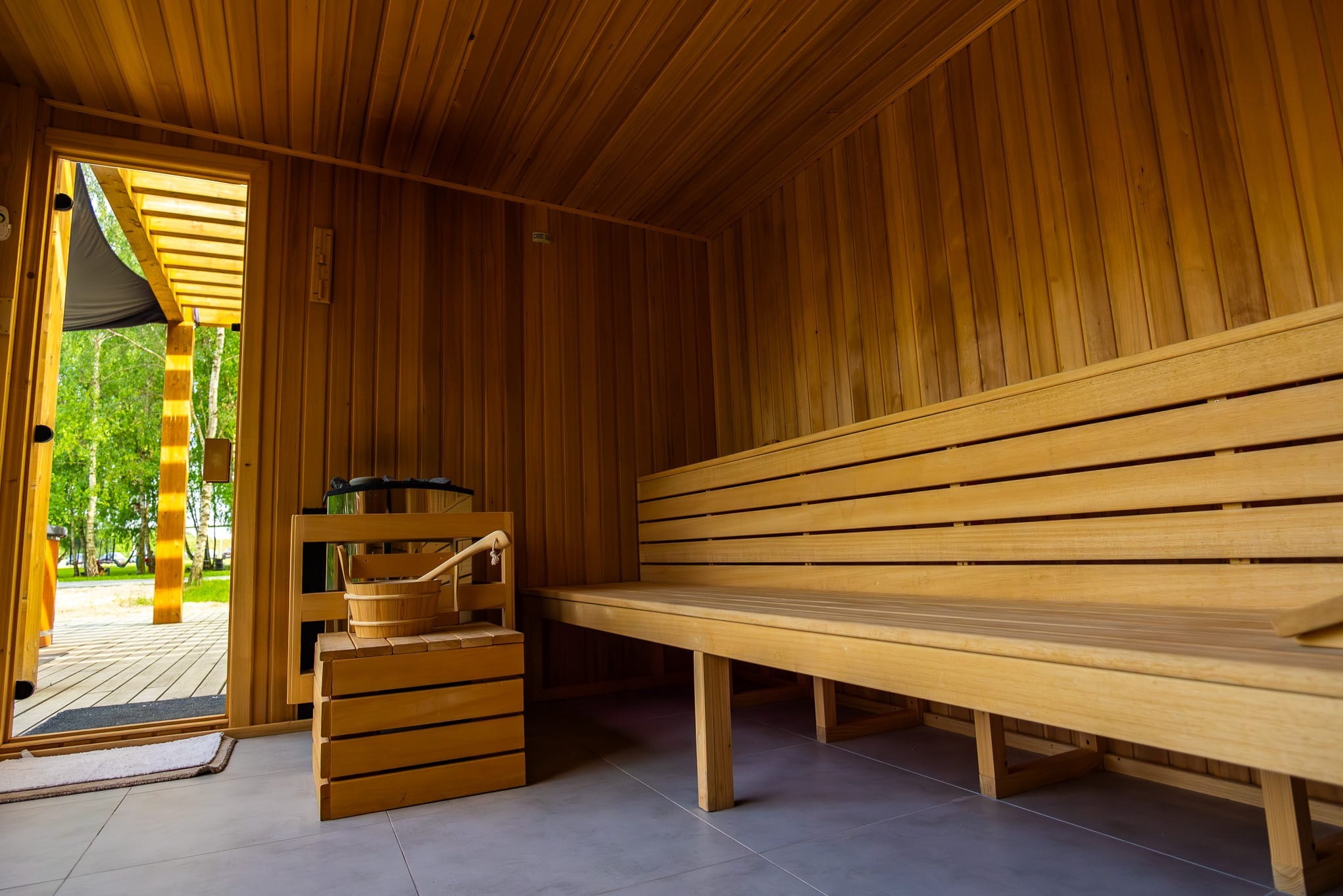 Wnętrze sauny z drewnianym wykończeniem, ławami i widokiem na zewnątrz przez otwarte drzwi.