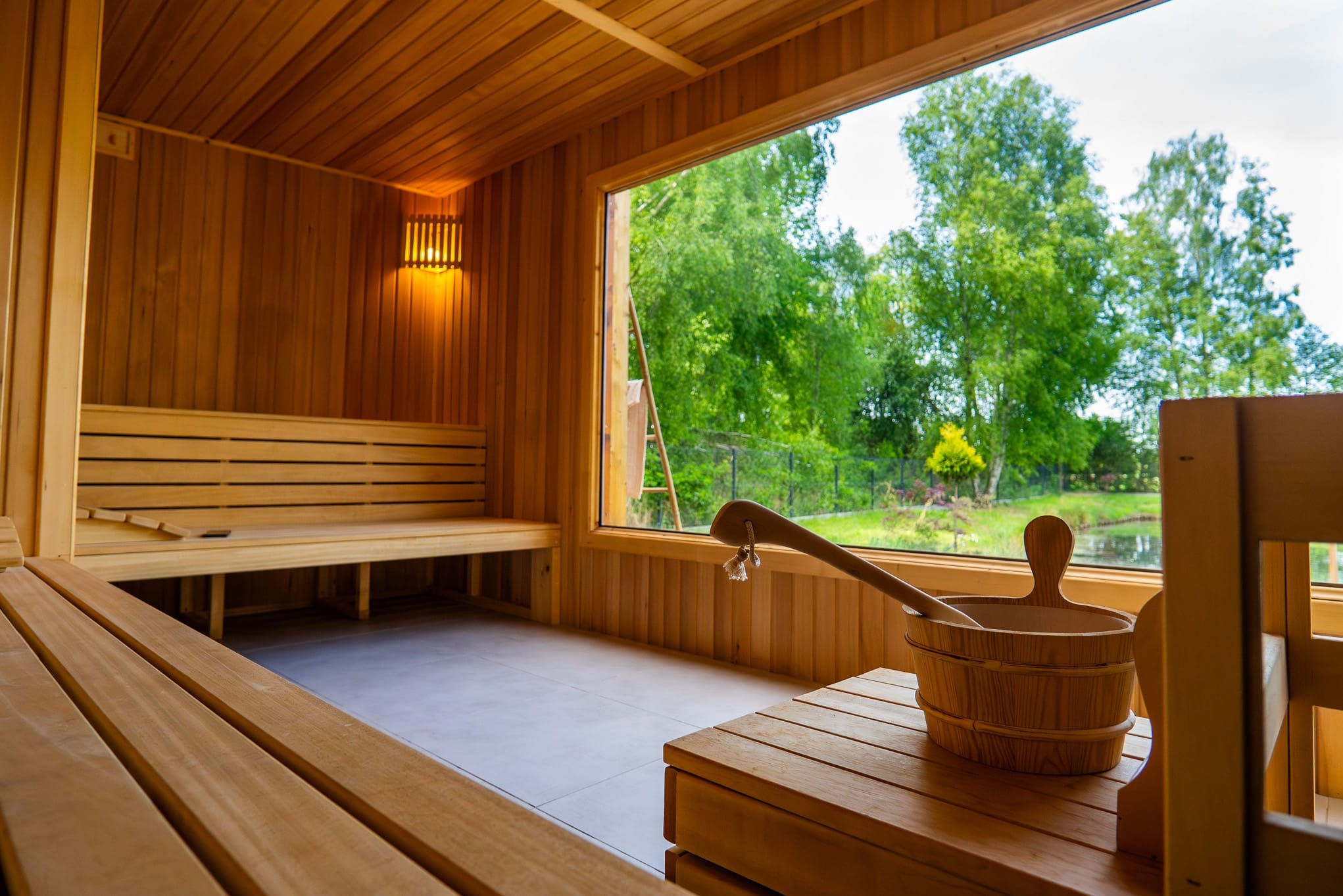 Wnętrze sauny wyłożone drewnem z widokiem na zieleń przez duże okno, drewniane ławy i wiadro z chochlą.