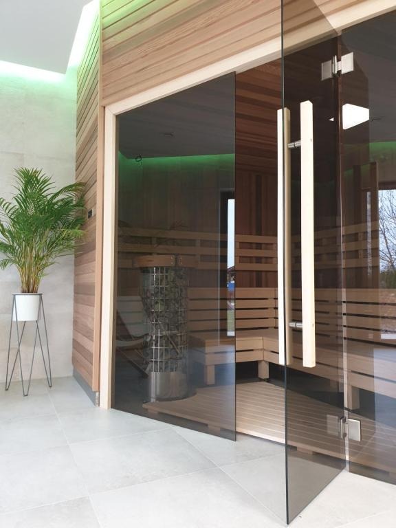 Nowoczesna sauna z drewnianym wykończeniem, przeszklonymi drzwiami z podłużnym uchwytem i metalowym piecem wypełnionym kamieniami. Obok wejścia stoi roślina doniczkowa w białej osłonce na metalowym...
