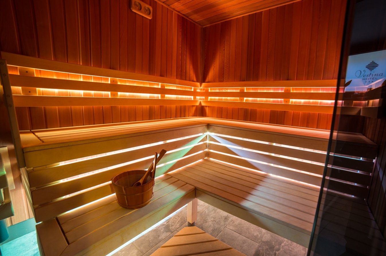 Wnętrze sauny z drewnianymi ławkami podświetlonymi ledami, wiaderko z chochelkami w rogu, widoczny fragment logo Vestina Hotel za szybą.