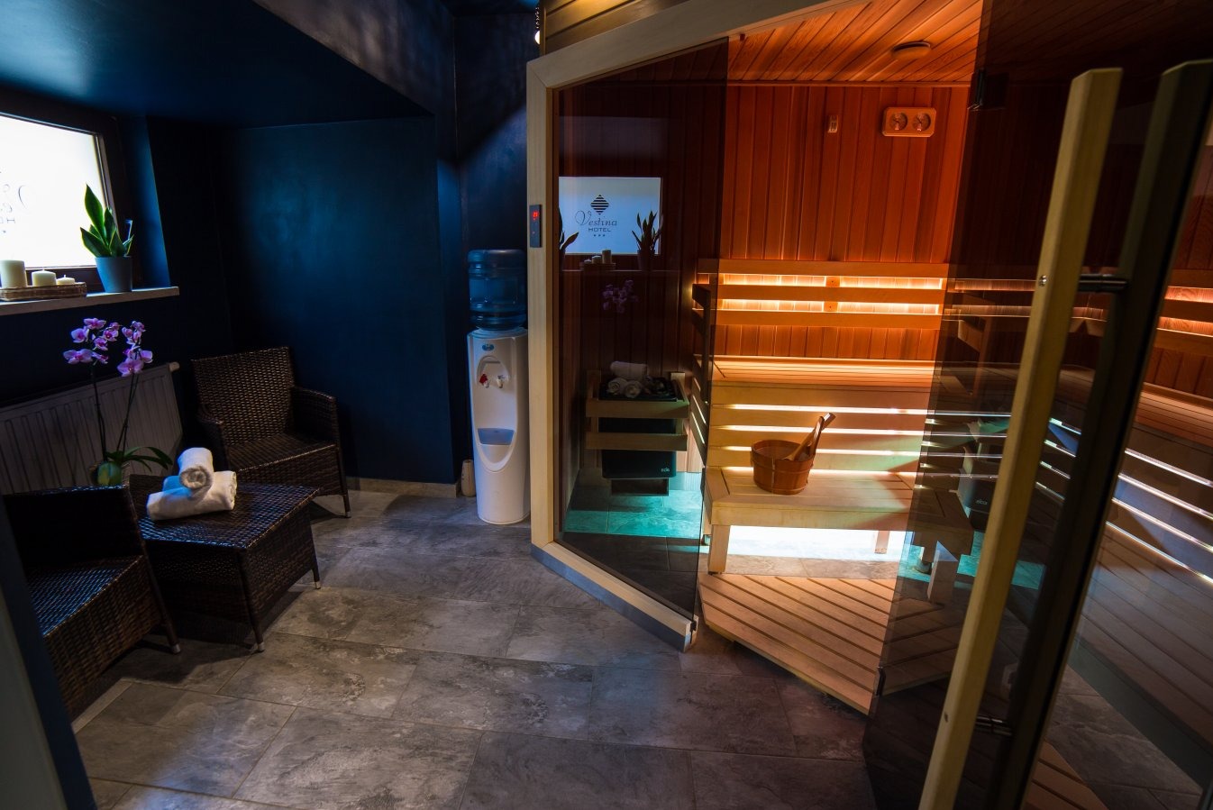 Wnętrze sauny z drewnianymi ławkami i oświetleniem, widok z zewnątrz przez szklane drzwi, obok strefa relaksu z rattanowymi fotelami, stolikiem, kwiatami i dystrybutorem wody.