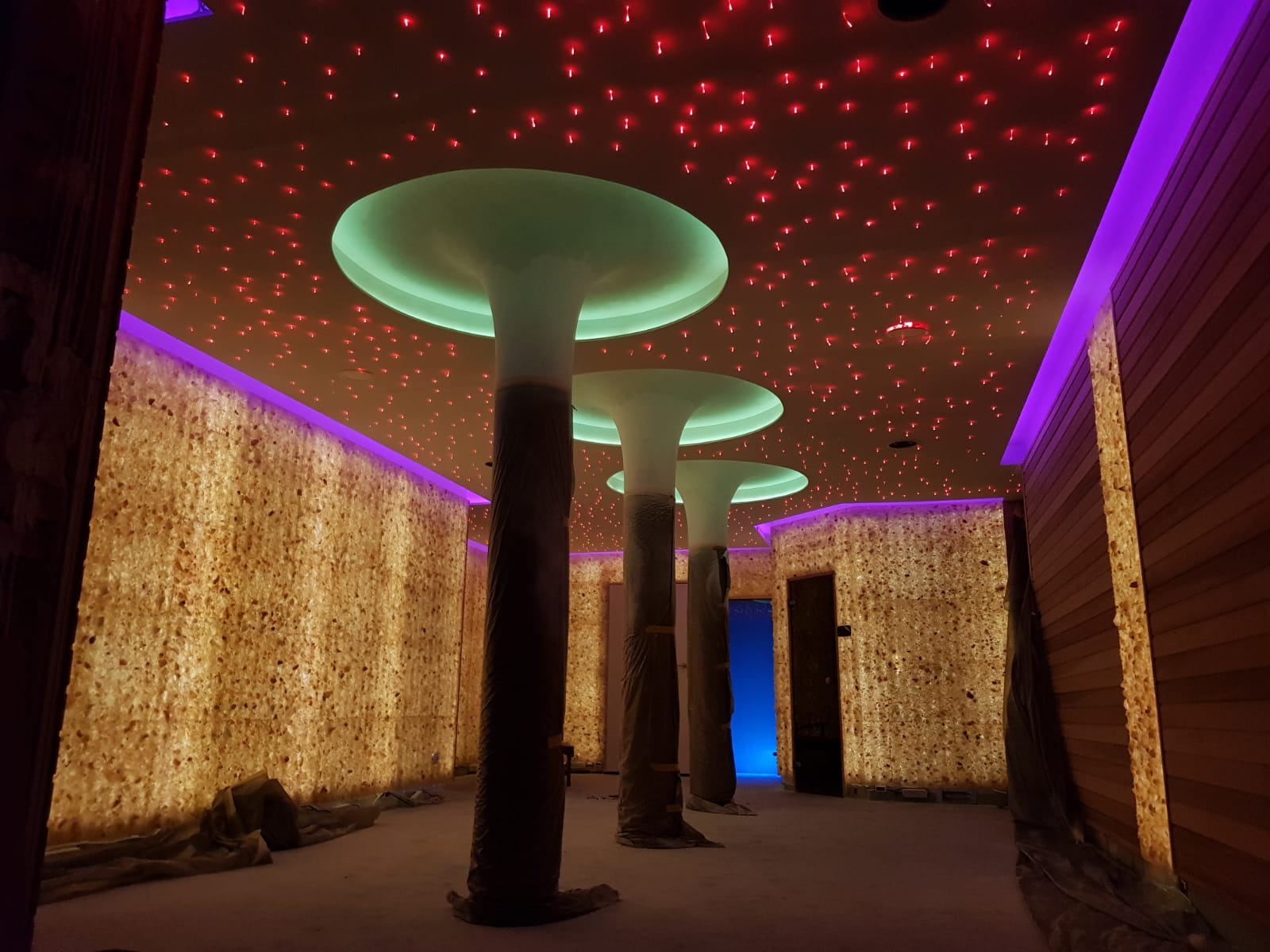 Wnętrze sauny z podświetlanymi ścianami z kamienia solnego, dekoracyjnym oświetleniem sufitowym w postaci czerwonych punktów i kolumnami z zielonymi kapitelami.