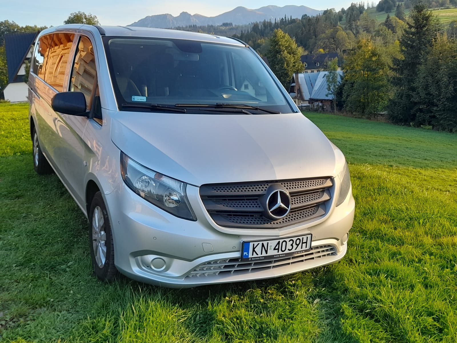 Srebrny Mercedes Vito zaparkowany na zielonej trawie z widokiem na góry i drewniane domy w tle, tablica rejestracyjna KN 4039H.