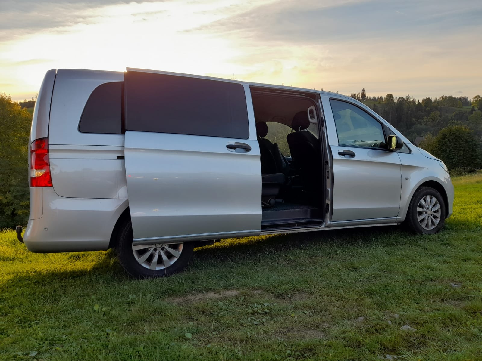 Srebrny van z otwartymi przesuwnymi drzwiami, zaparkowany na trawie o zachodzie słońca; widoczne czarne fotele wewnątrz.