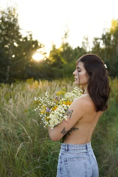 Portret kobiety z odsłoniętymi plecami, trzymającej bukiet polnych kwiatów na tle łąki o zachodzie słońca, z widocznym tatuażem na ramieniu i kwiatem we włosach.