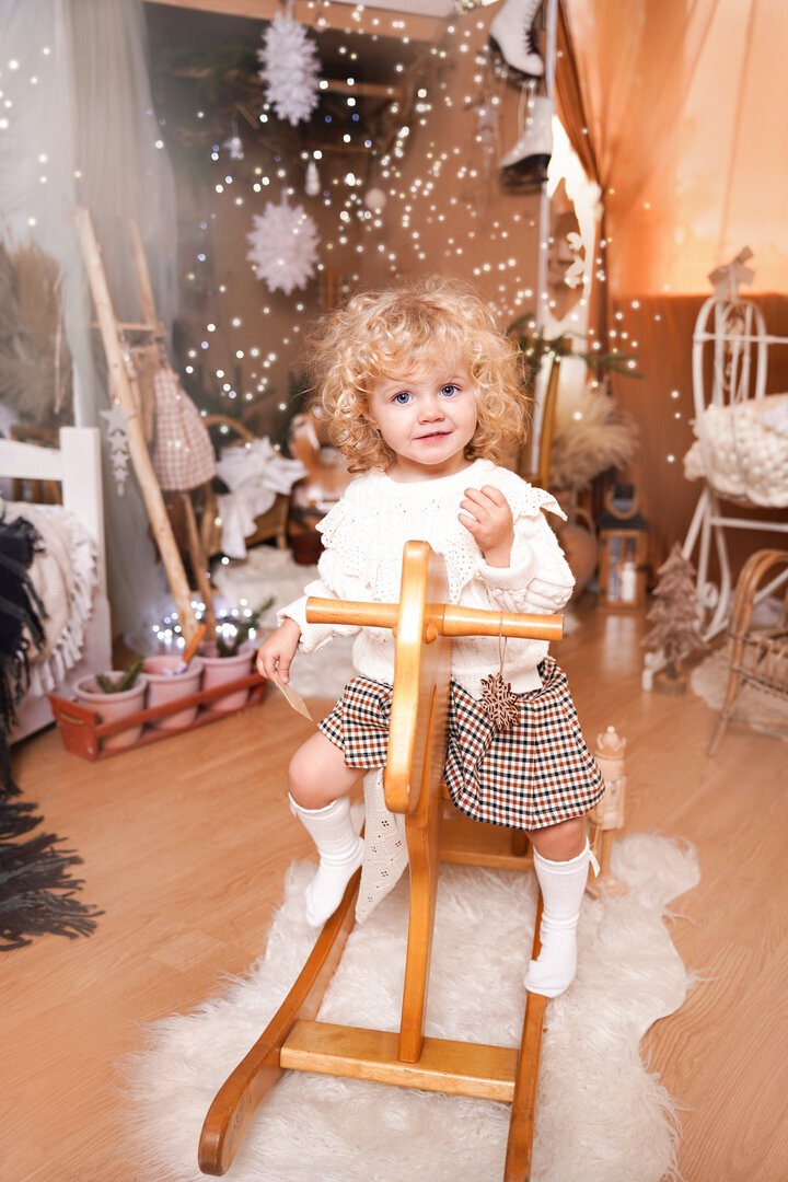 Uśmiechnięta dziewczynka z kręconymi włosami siedzi na drewnianym koniku na biegunach, w tle dekoracje świąteczne i lampki.