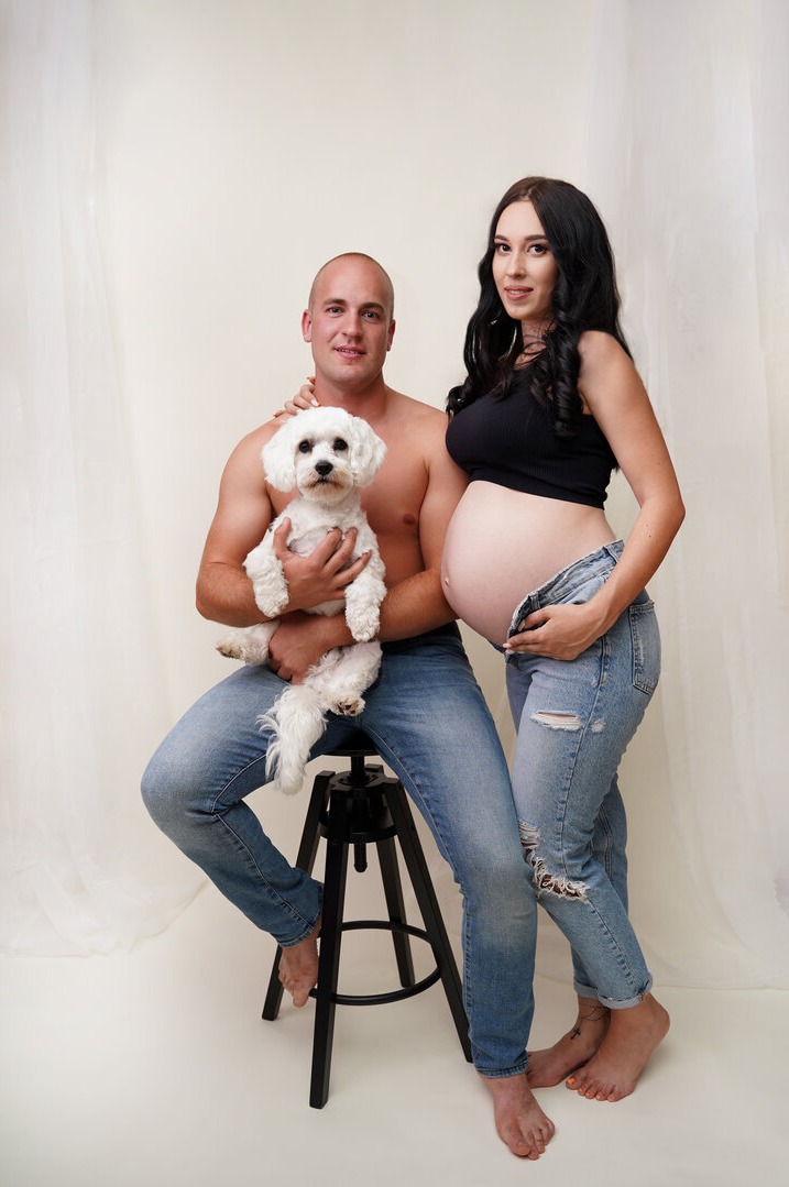 Przyszła mama w podartych jeansach i czarnym topie, pozuje z partnerem trzymającym białego maltańczyka na czarnym stołku barowym na tle kremowej zasłony.