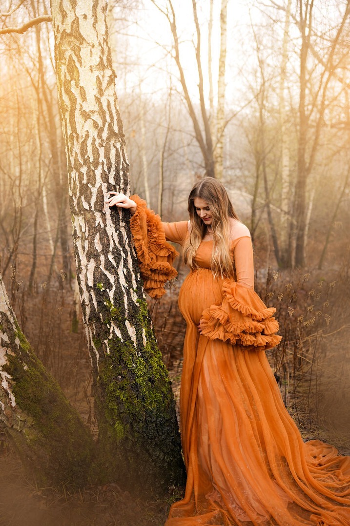 Kobieta w pomarańczowej, zwiewnej sukni ciążowej z falbanami pozuje opierając się o brzozę porośniętą mchem w lesie, z delikatnym oświetleniem słonecznym.
