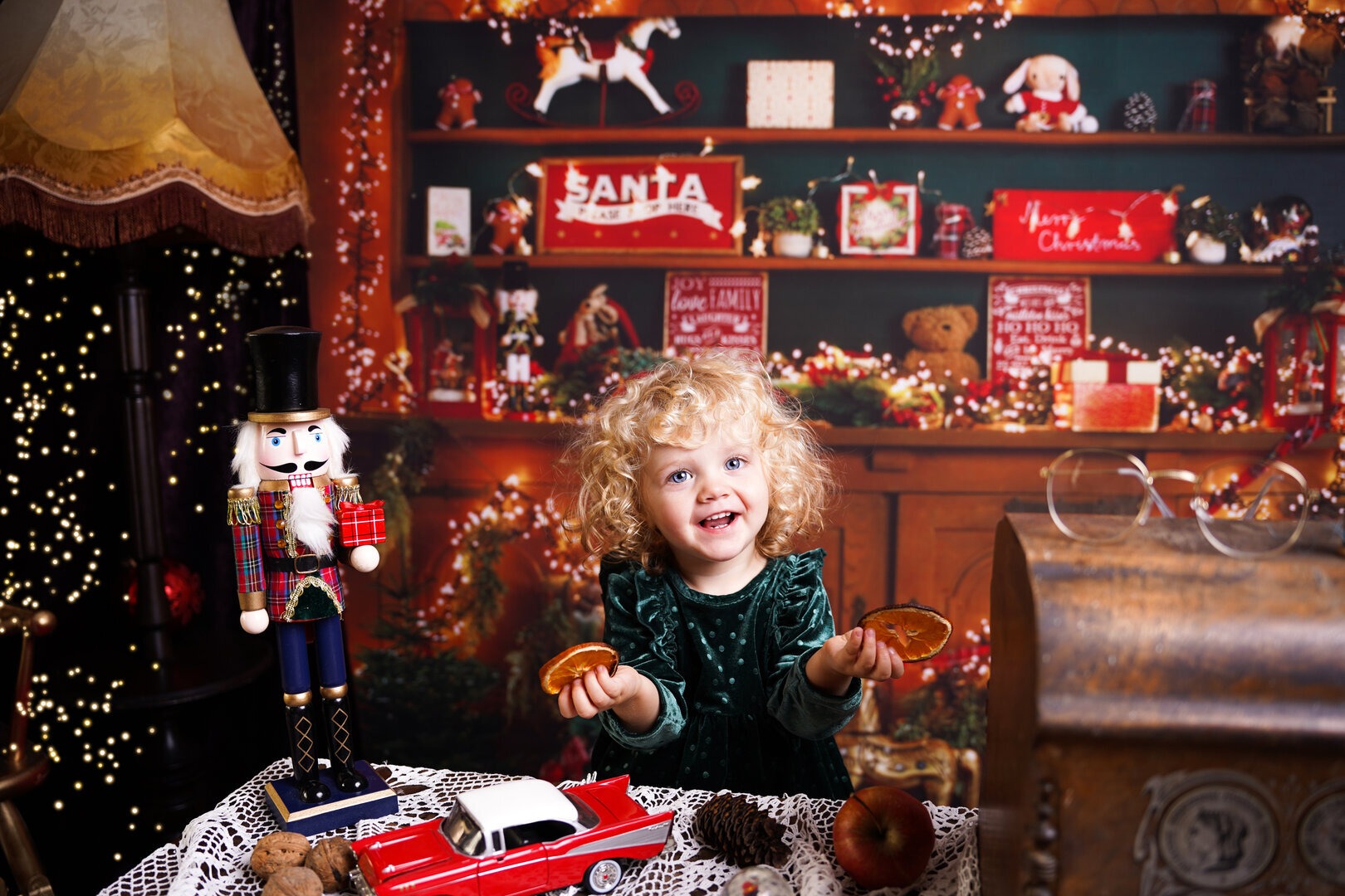Uśmiechnięta dziewczynka z kręconymi włosami w zielonej sukience trzyma plasterki pomarańczy na tle świątecznej dekoracji z dziadkiem do orzechów, zabawkami i światełkami.