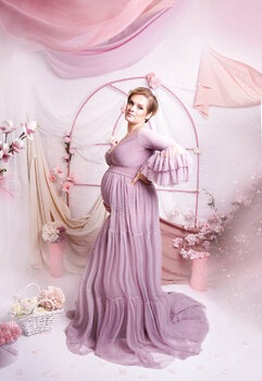 Kobieta w długiej, liliowej sukni ciążowej, pozująca na tle dekoracji z kwiatów i tkanin w pastelowych odcieniach różu i bieli. Ręka oparta na biodrze, podkreślony brzuch, delikatny uśmiech.