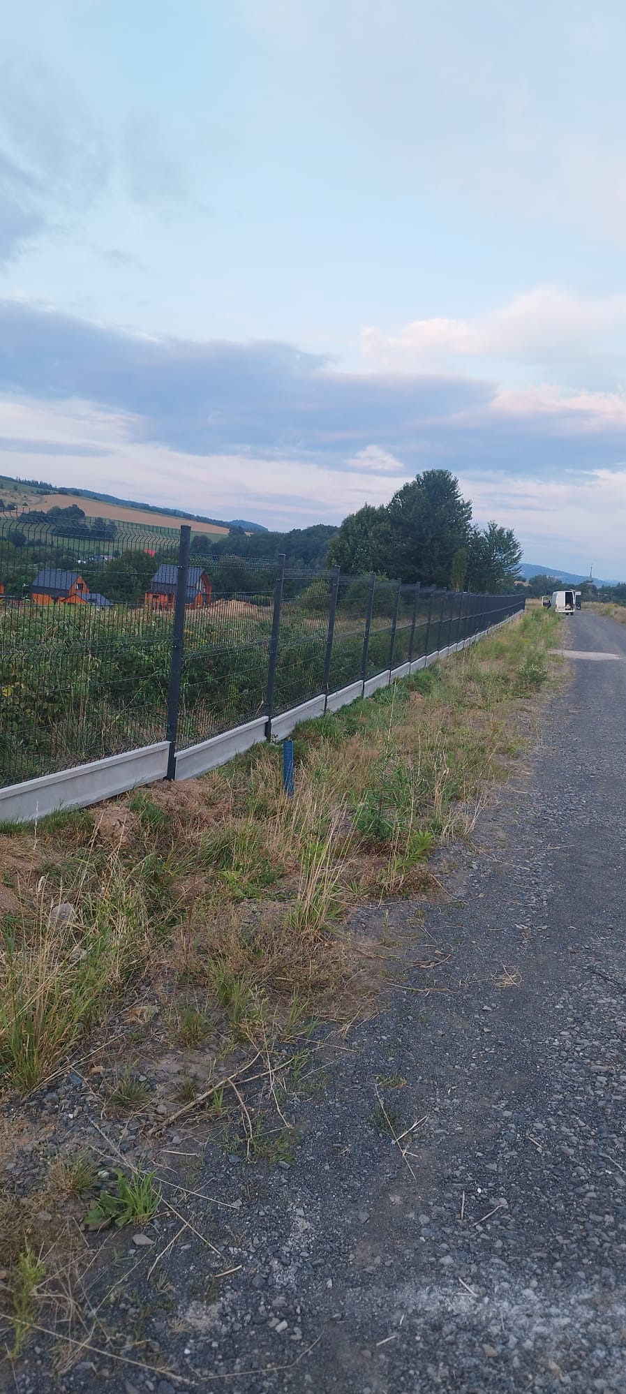 Długi, ciemny płot panelowy z podmurówką betonową, ciągnący się wzdłuż polnej drogi, z widokiem na pagórkowaty krajobraz i zachmurzone niebo w tle.