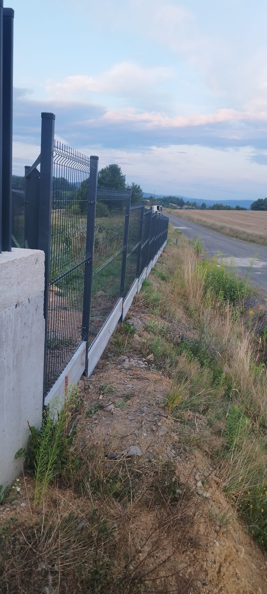 Ciemnoszare ogrodzenie panelowe z podmurówką betonową w Wisznia Mała, widok z boku na tle polnej drogi i pól uprawnych.