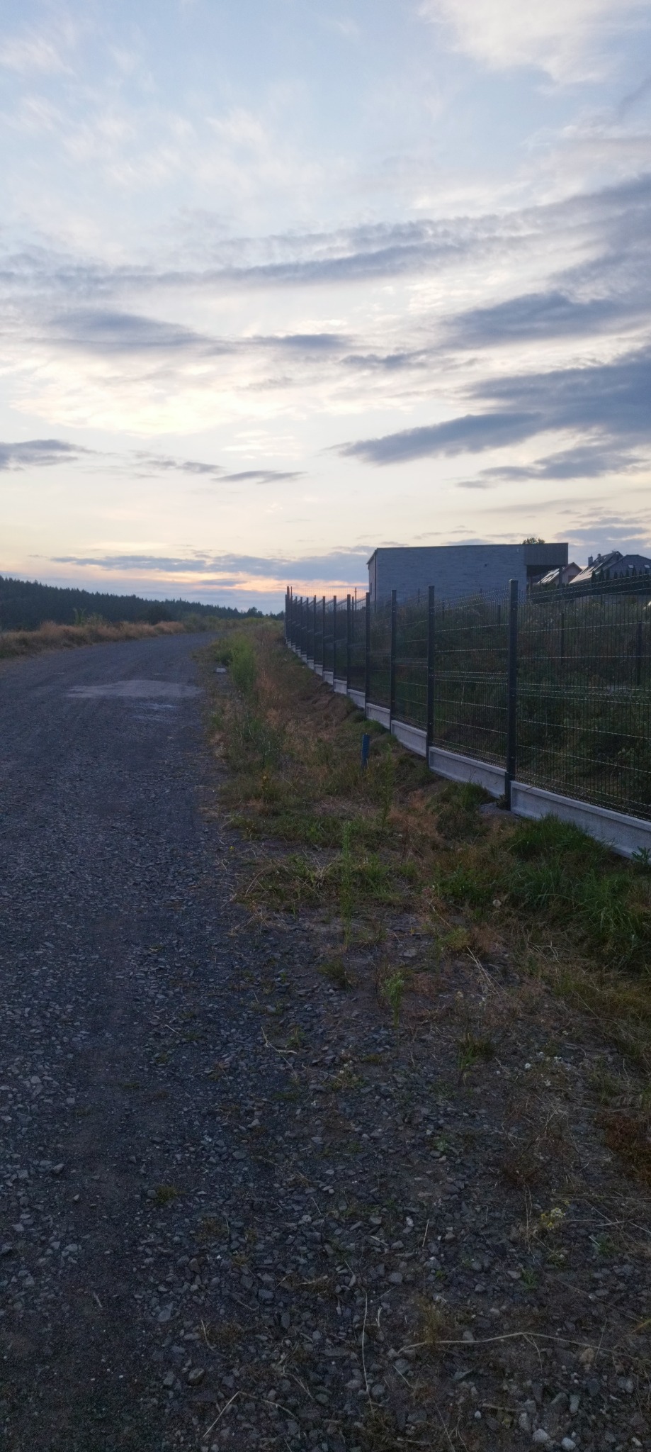 Długa linia ogrodzenia panelowego z podmurówką betonową, widoczna z perspektywy drogi gruntowej, w tle budynek gospodarczy i zachodzące słońce.