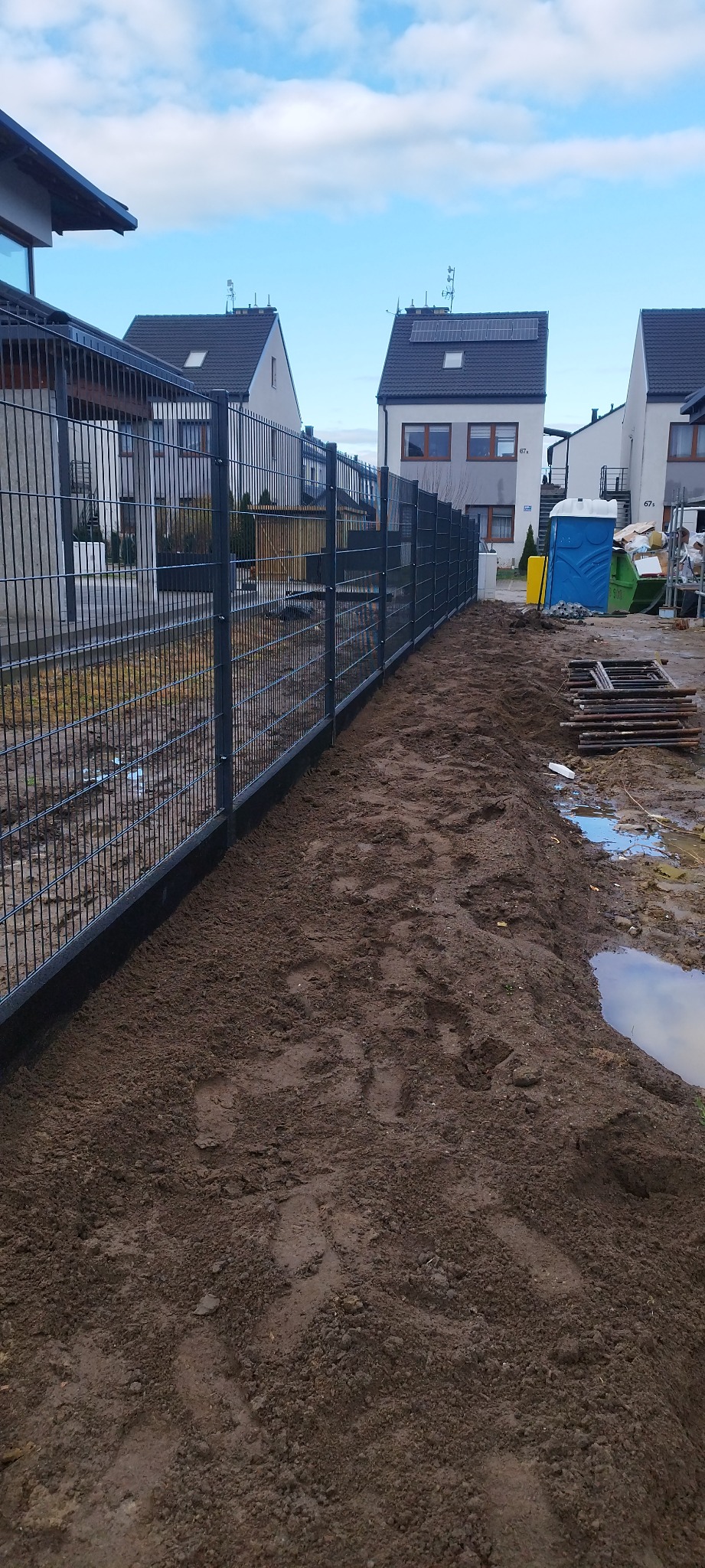 Długi, szary panelowy płot z widocznymi słupkami, ciągnący się wzdłuż granicy działki, z ziemią świeżo obsypaną u podstawy, na tle domów jednorodzinnych i błękitnego nieba.