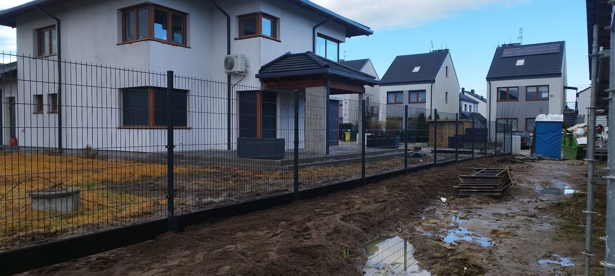 Częściowo ukończona instalacja czarnego ogrodzenia panelowego na posesji z nowym domem, widoczna wykopana ziemia i kałuże.