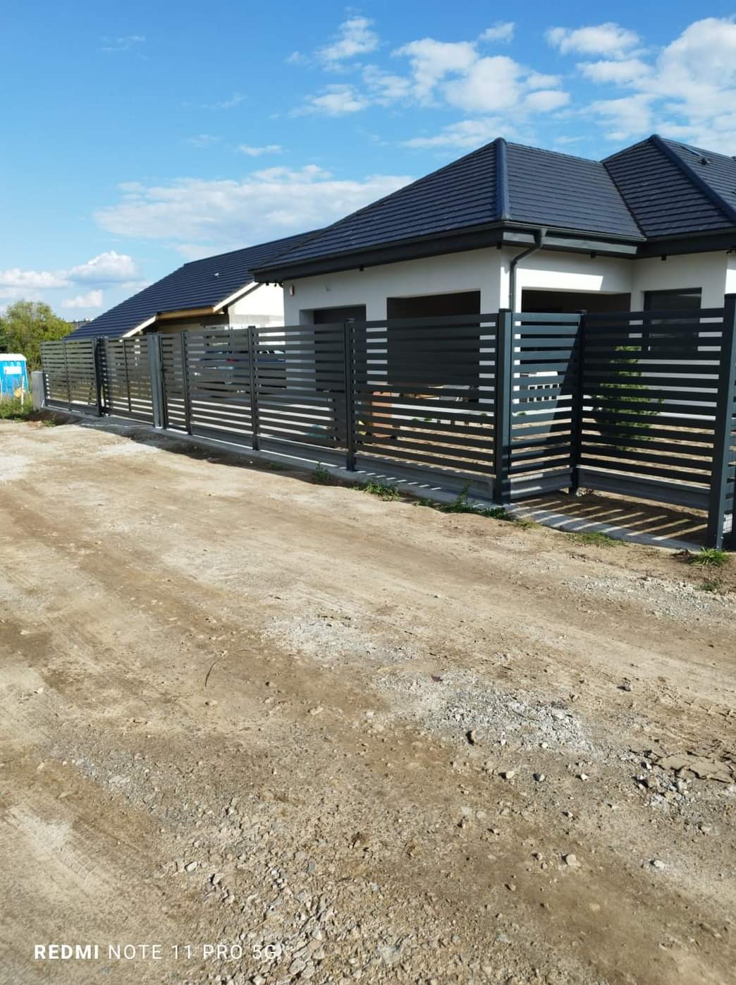 Nowoczesne ogrodzenie panelowe z poziomych, szarych profili, dopasowane kolorystycznie do dachu domu, na tle błękitnego nieba z chmurami.