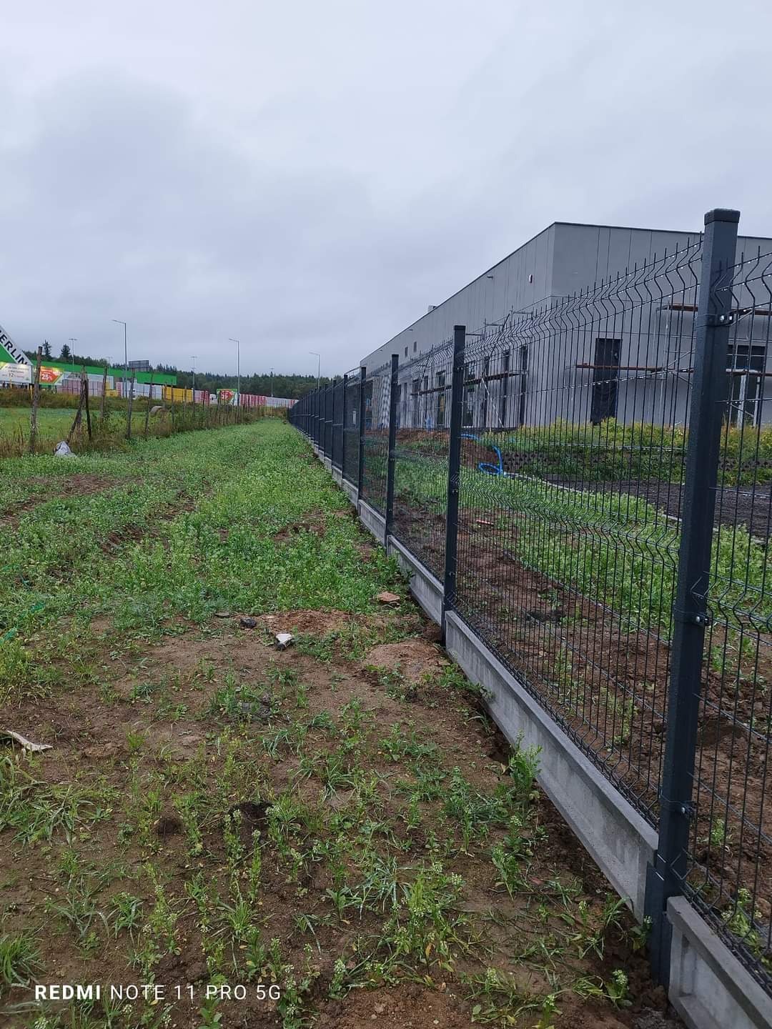 Długi, szary panelowy płot z betonową podmurówką, ciągnący się wzdłuż terenu przemysłowego z budynkiem w tle, widok z poziomu gruntu na zewnątrz ogrodzonego obszaru.