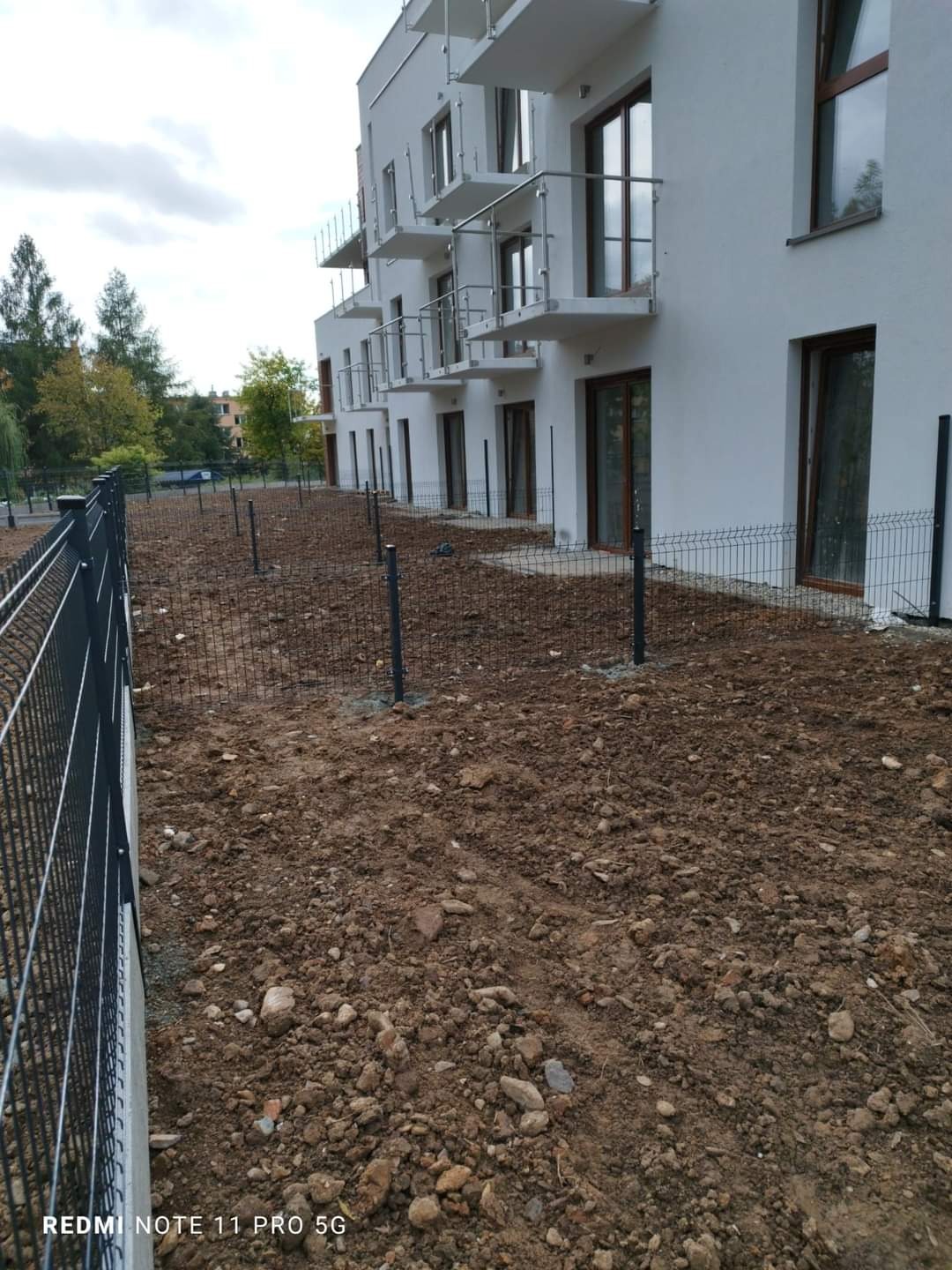 W trakcie montażu ogrodzenia panelowego wokół nowego budynku mieszkalnego, widoczne słupki i częściowo zamontowane panele na nierównym, przygotowanym terenie.