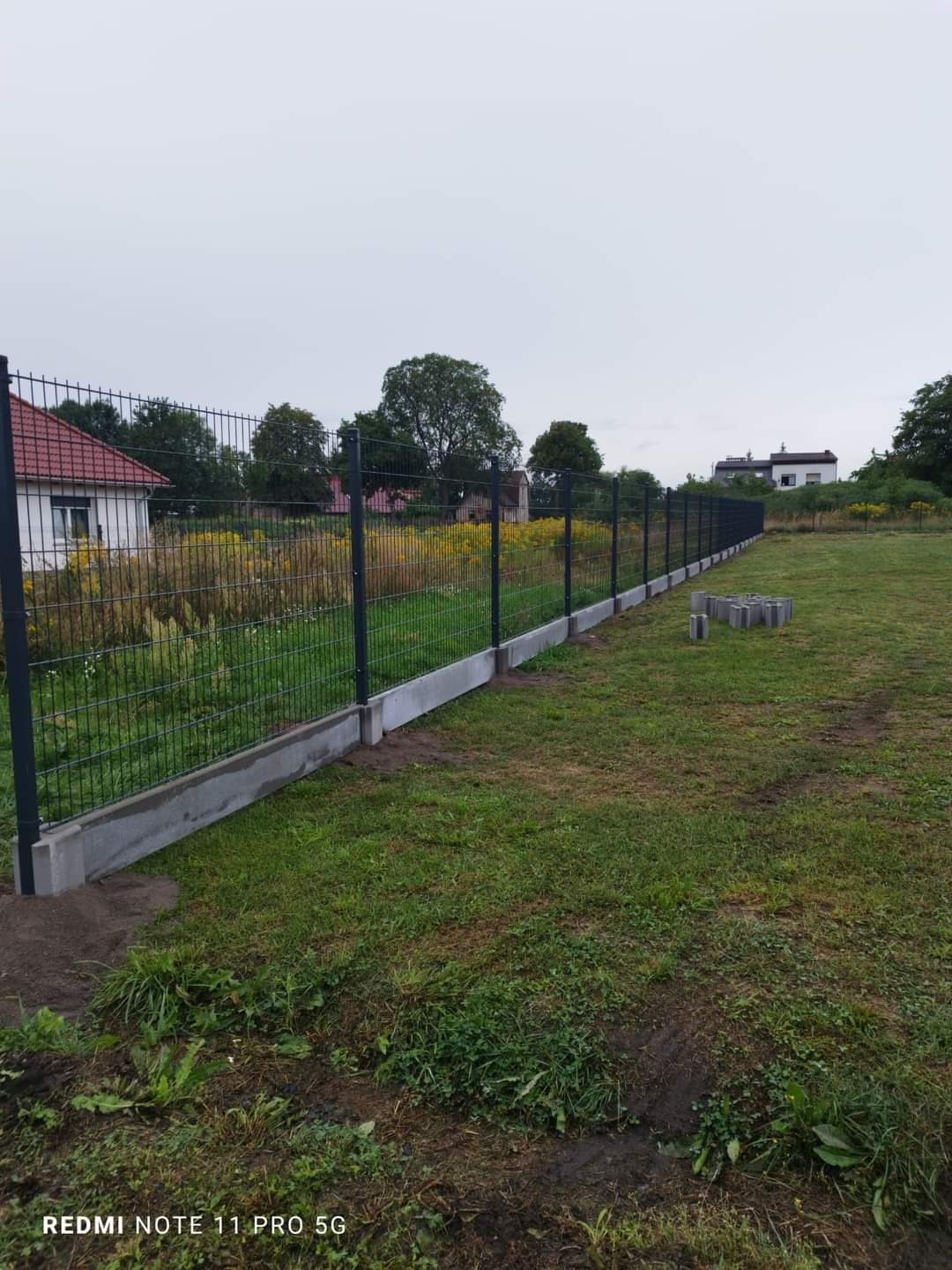 Nowo postawione ogrodzenie panelowe z podmurówką betonową na działce z widokiem na domy w tle, kilka betonowych bloczków leży obok ogrodzenia.