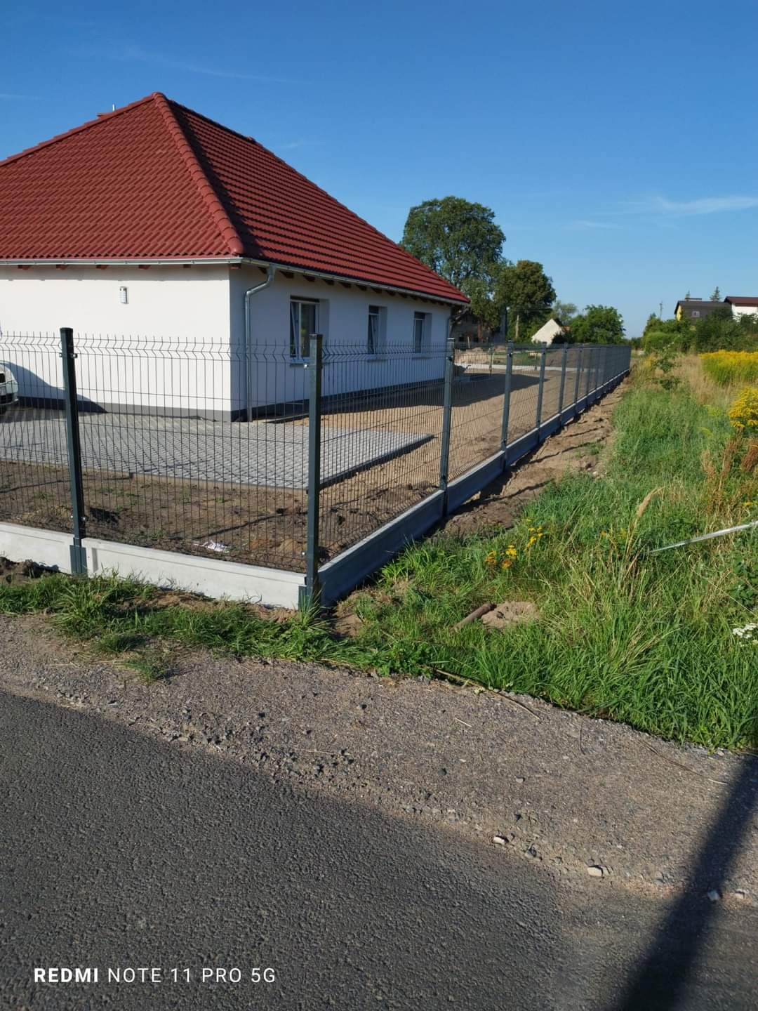 Nowo zainstalowane ogrodzenie panelowe z podmurówką betonową, widok z drogi asfaltowej, dom z czerwoną dachówką w tle.
