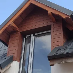 Fragment elewacji domu z oknem w szczycie dachu, obłożonym brązową boazerią, z widoczną czarną dachówką i elementami obróbek blacharskich.