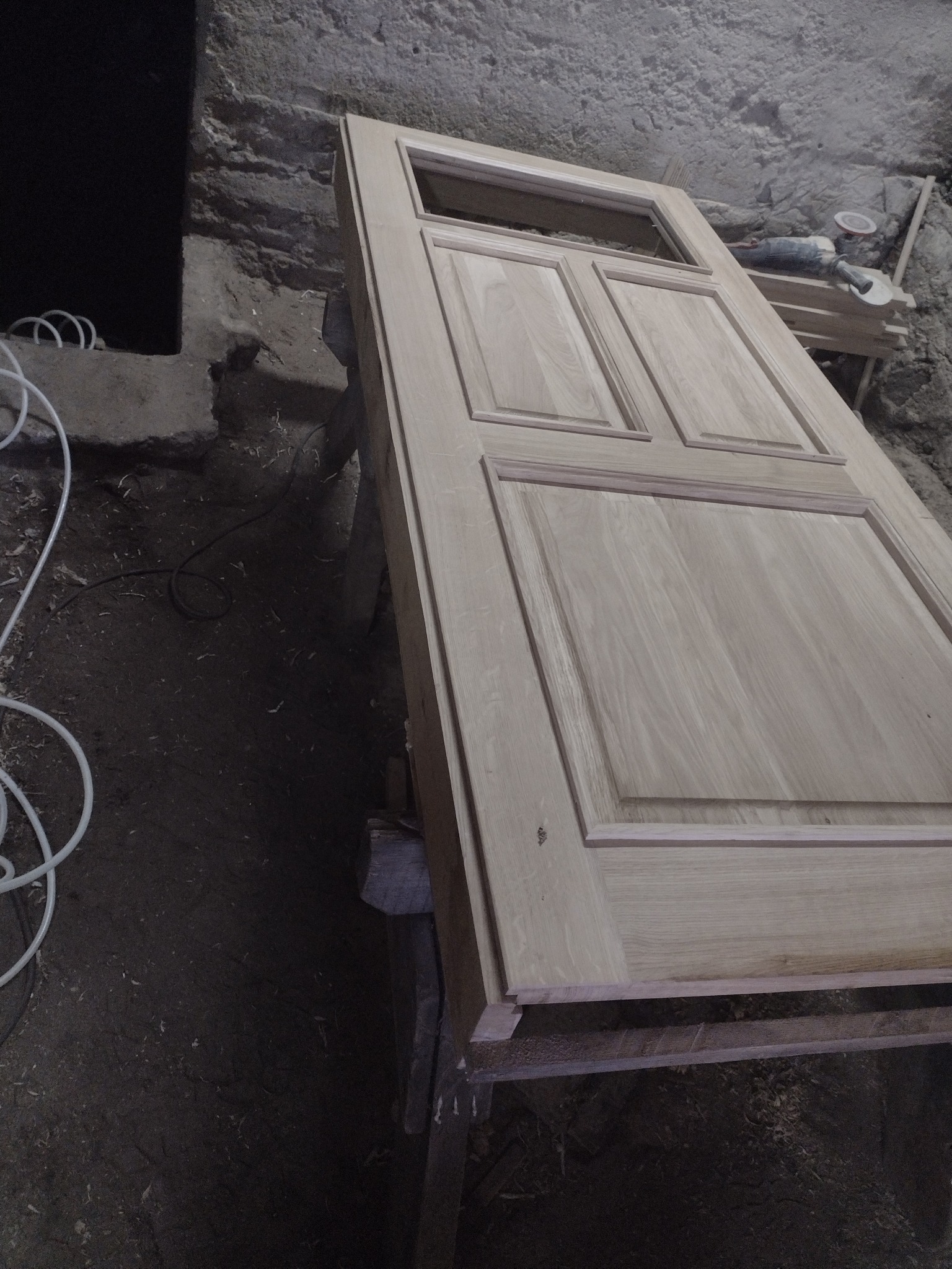 Surowa, drewniana rama dachu z widocznymi panelami, leżąca na drewnianych kozłach w surowym, warsztatowym otoczeniu z betonową ścianą w tle.